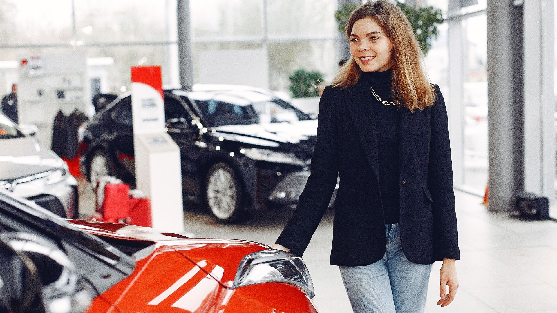 Happy stylish young female in jeans and black jacket standing near modern red shiny car in contemporary car showroom and looking at purchase dreamily