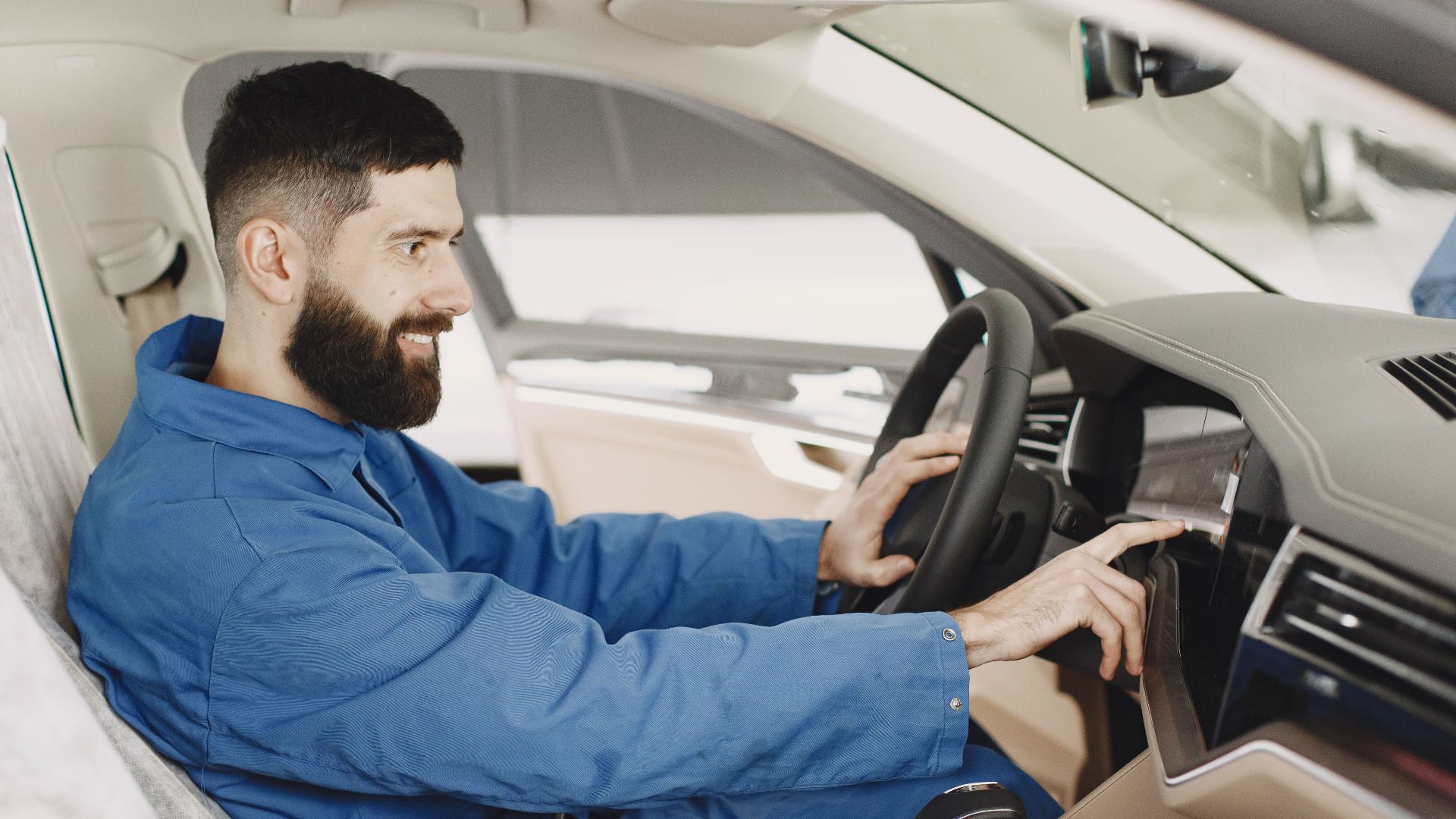 Mechanic in blue coveralls interacts with car dashboard, smiling and focused.