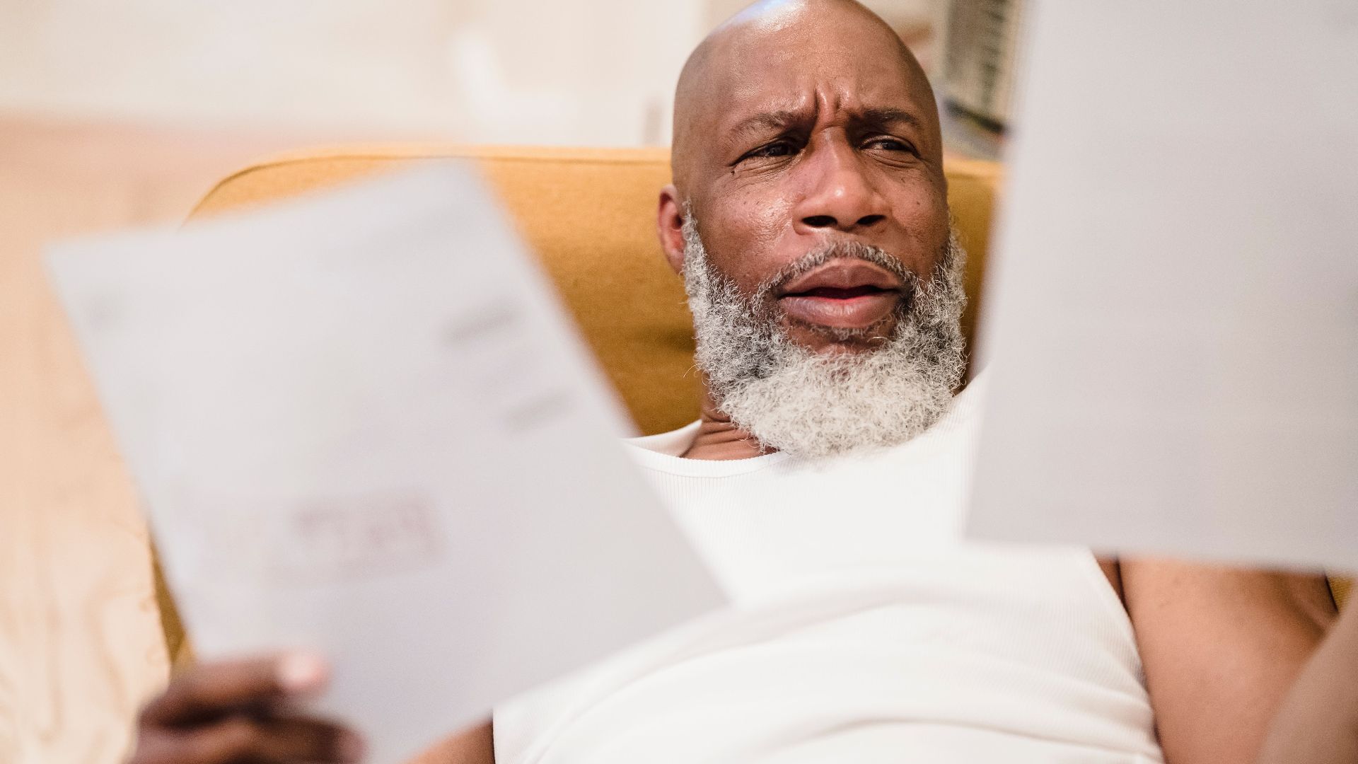 A middle-aged man looks concerned as he reviews household bills, sitting in a comfortable chair indoors.