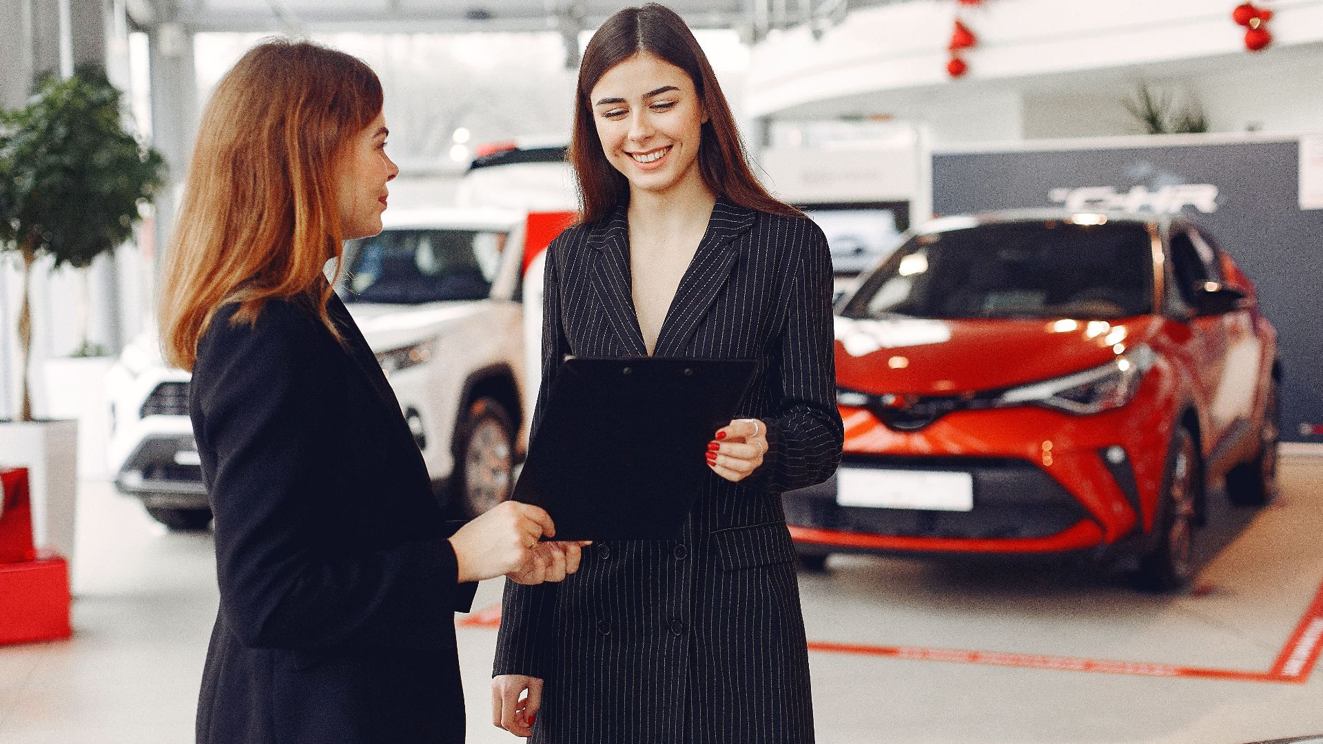 Cheerful young friendly dealer in formal stylish black dress showing contract to smiling female customer in black jacket while standing in car showroom against new shiny automobiles