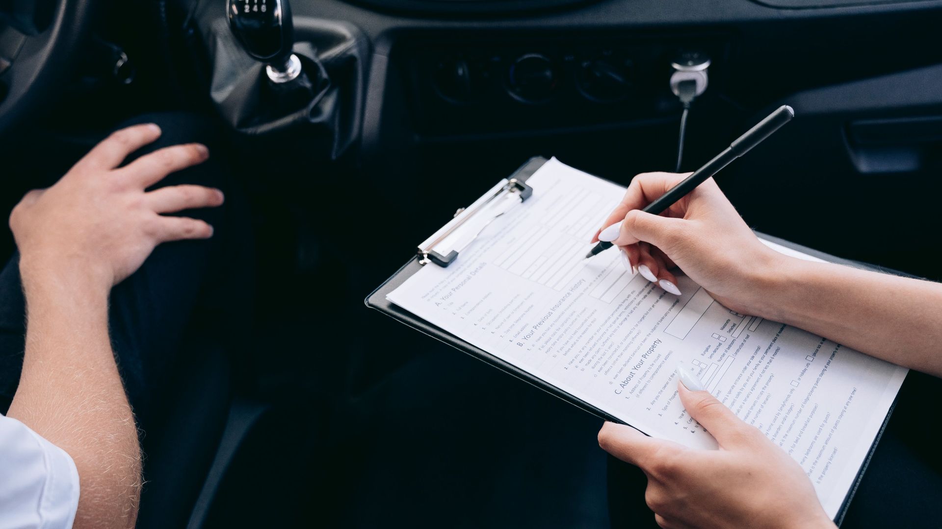 Close-up of a person writing on a clipboard inside a car, showing hands and a gear shift.