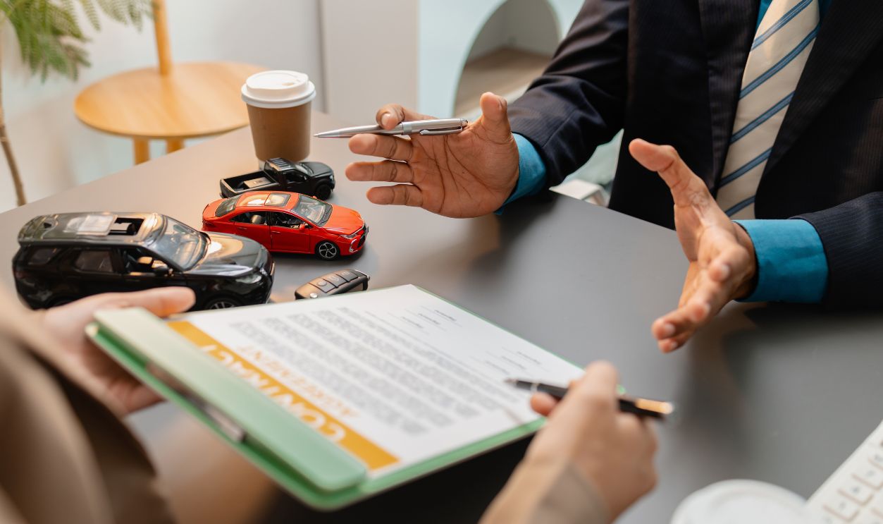 A business meeting between a car dealer and a client discussing vehicle purchase agreement. The contract is being reviewed and explained before signing, representing trust and cooperation.