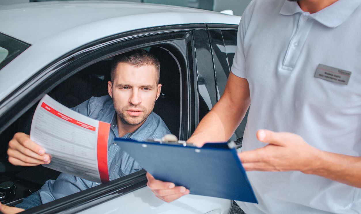 Seller stands at white car and holds tablet in hands. He point on it. Another man sits in car and holds insurance paper. He is serious and concentrated. They have conversation.