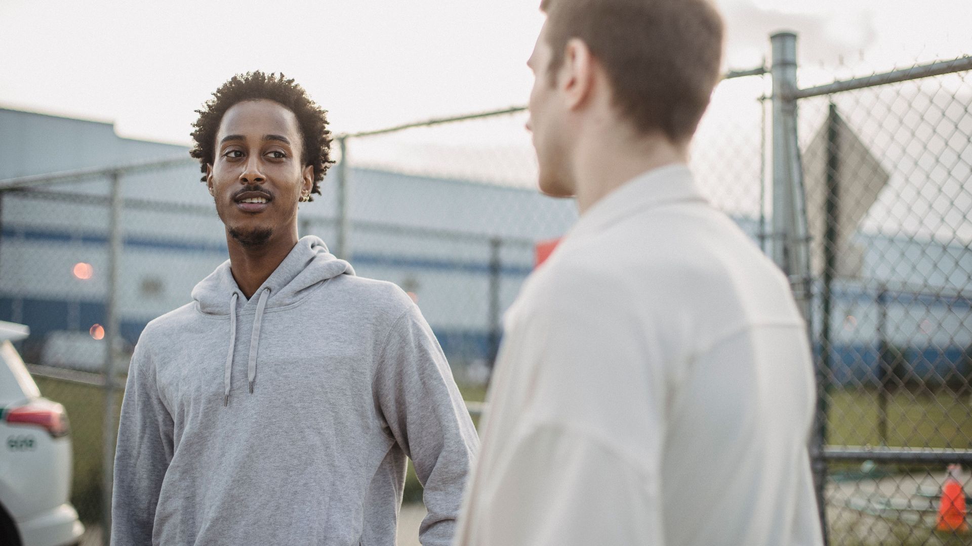 African American male standing with friend against metal grid near car on parking lot
