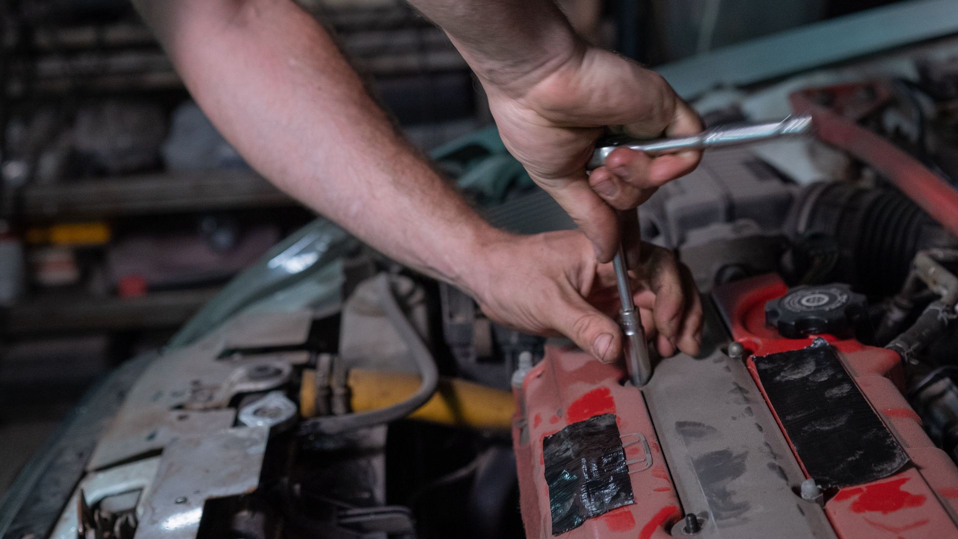 Mechanic repairs car engine in a dimly lit auto garage, focused on fixing the vehicle.