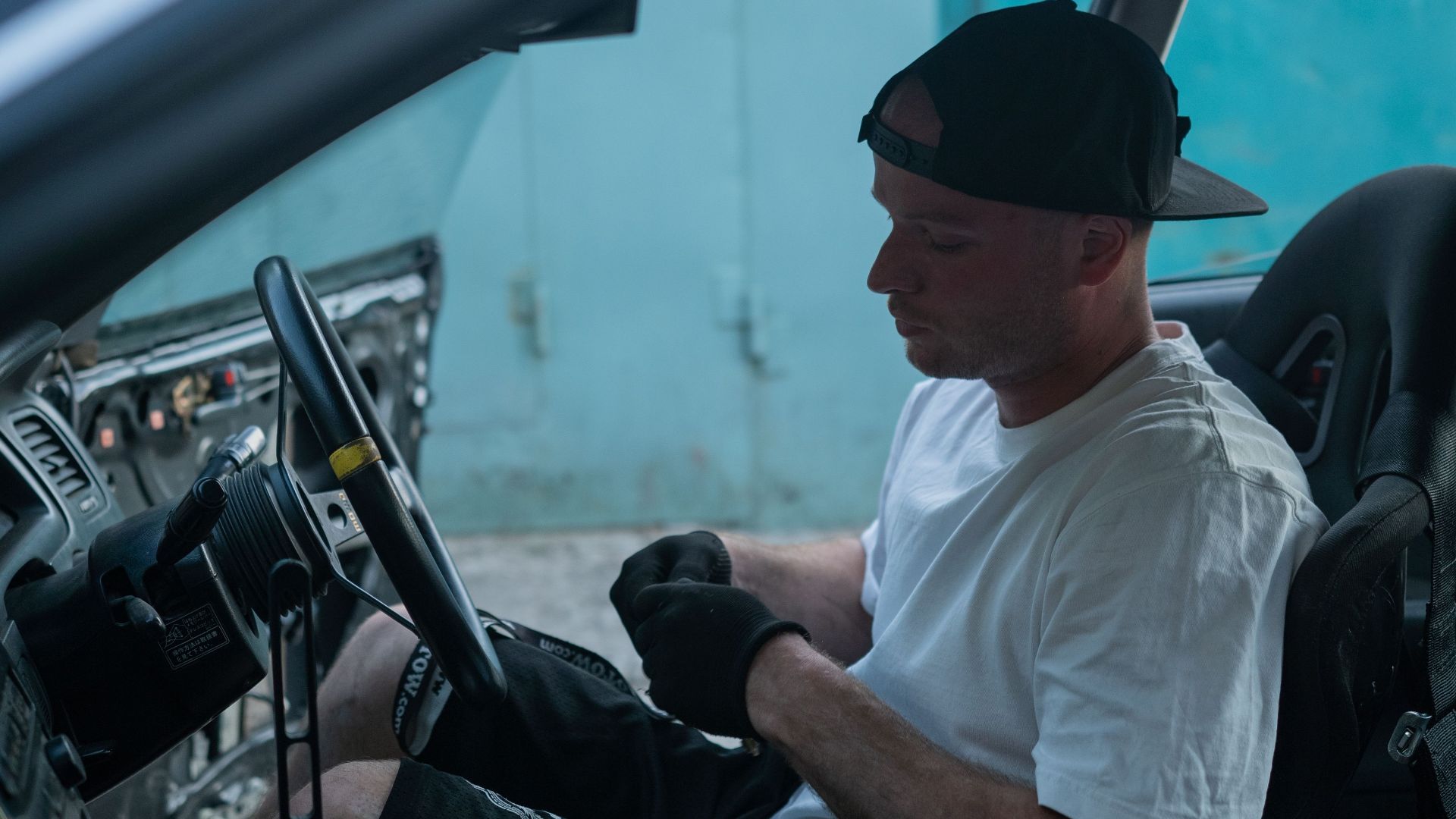 A mechanic in a black cap and gloves adjusts the steering wheel inside a car.