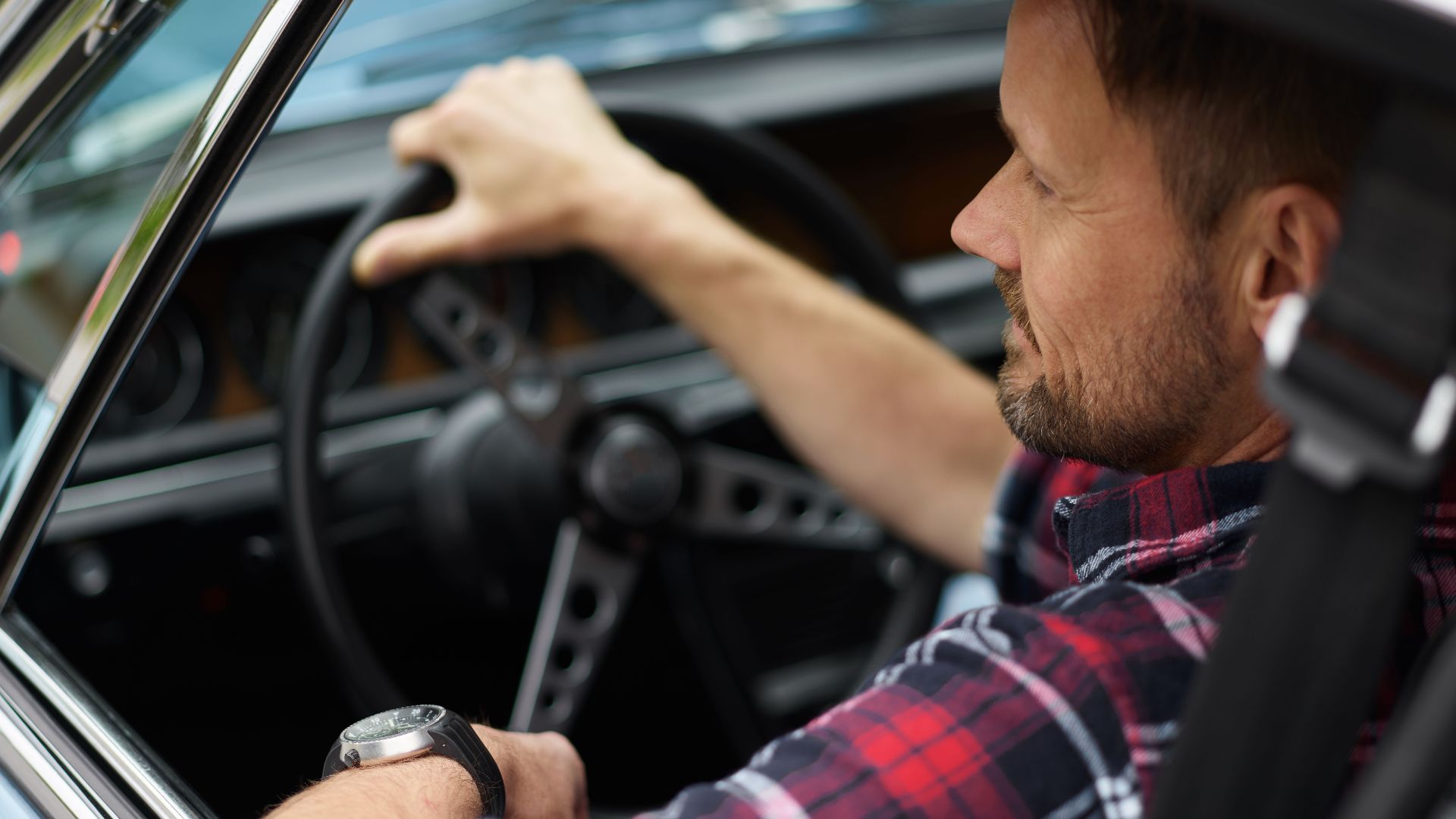 From above back view of confident male with short beard and mustache in plaid shirt sitting in automobile with hand on steering wheel