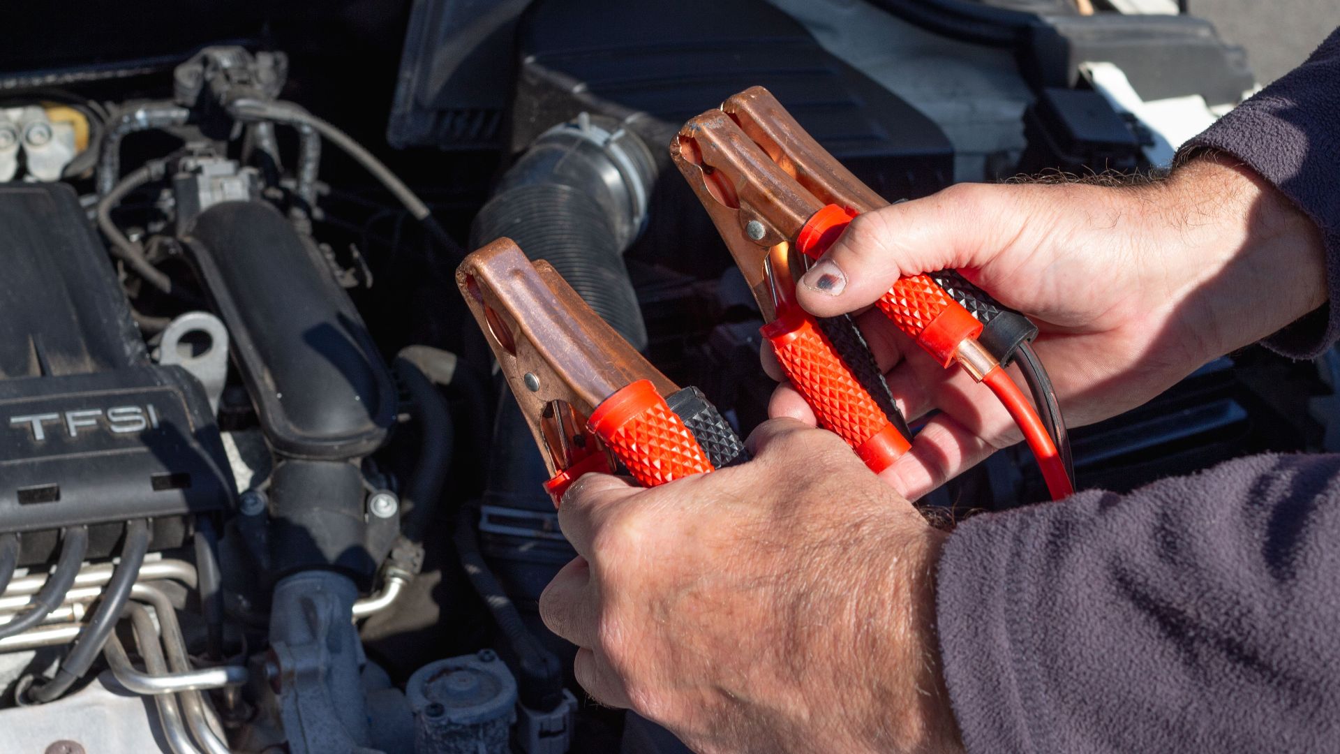 Close-up of hands holding jumper cables near a car engine, ready for repair.