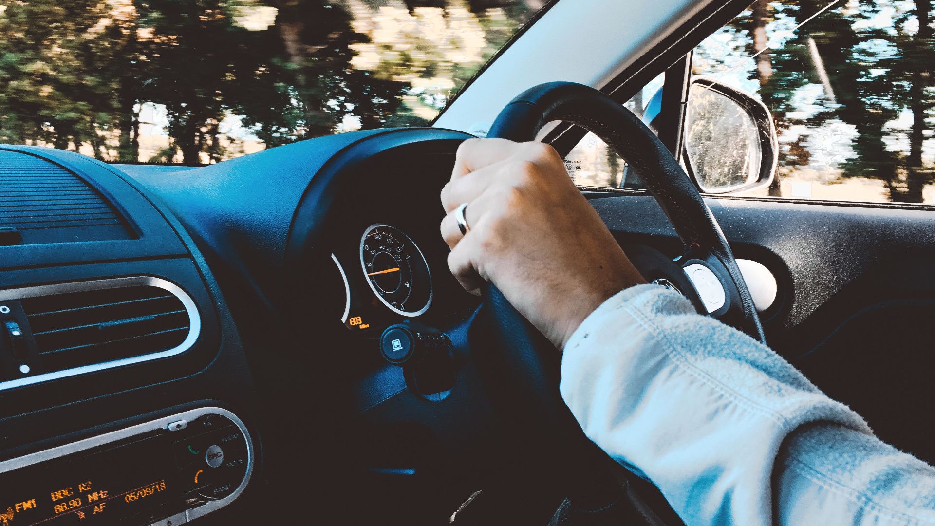 Driver's hand on steering wheel of a modern car during a daytime drive through scenic outdoors.