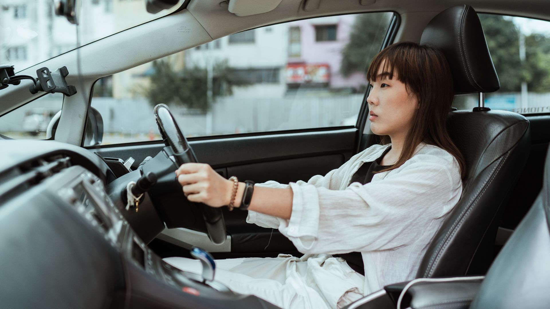 Side view unemotional Asian female in casual white clothes steering right hand drive car along urban streets in daytime