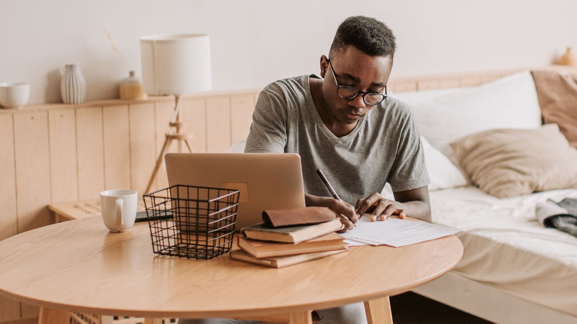 Man sitting barefoot at a table in a cozy bedroom, working on a laptop with notes.