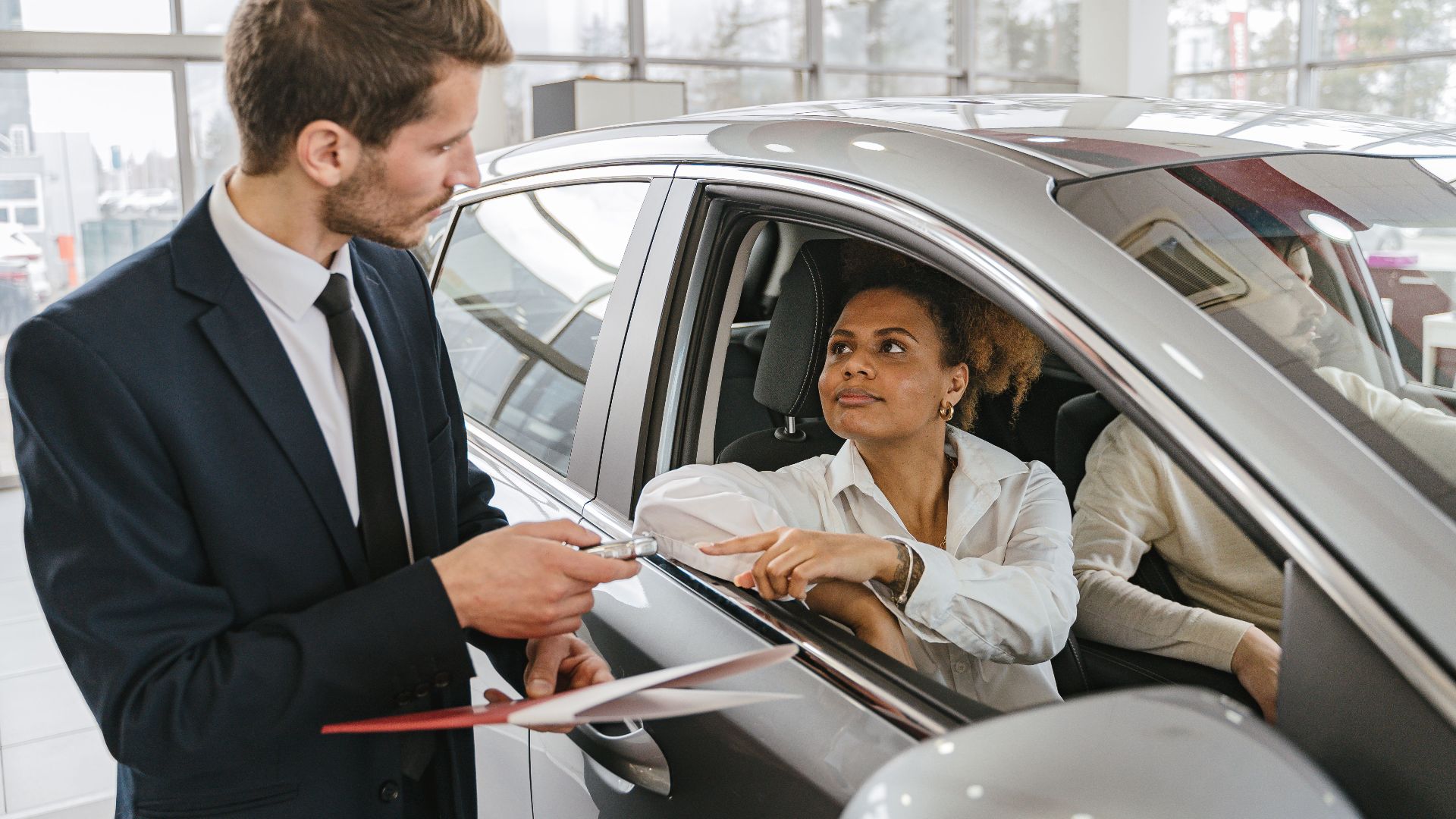 A car dealer hands keys to a woman sitting inside a vehicle in a car showroom.
