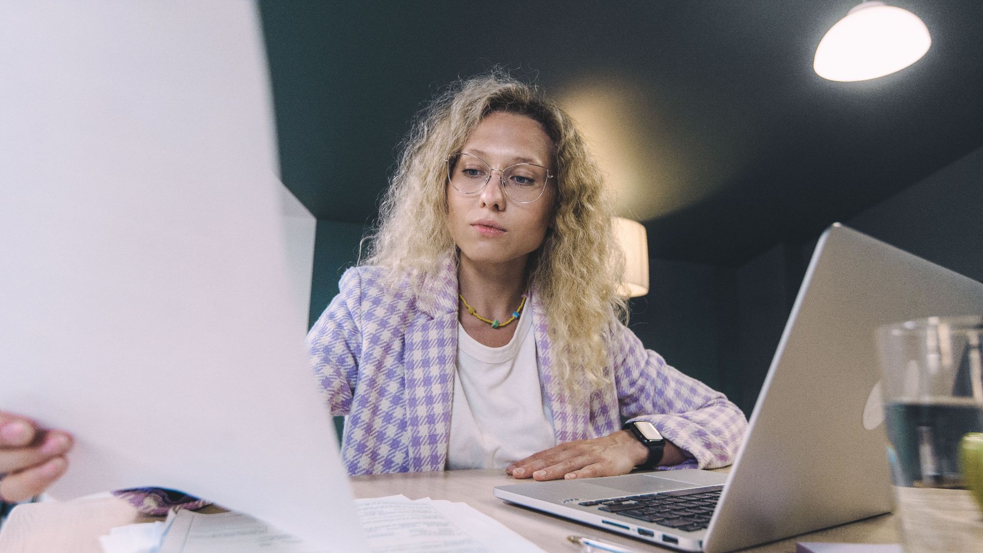 Woman in an office environment reviewing documents with focus, surrounded by technology.