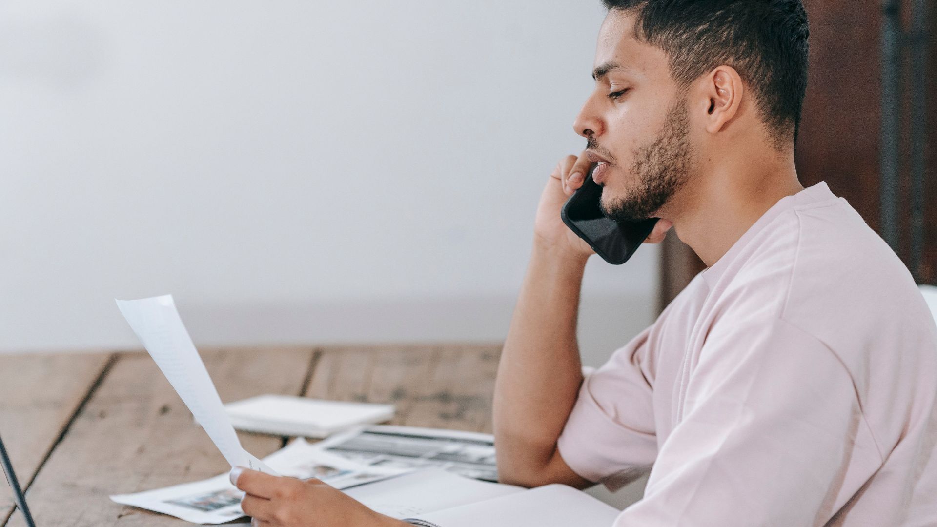 Side view of young Hispanic male remote employee discussing documents during phone call while working online with laptop and papers in home office