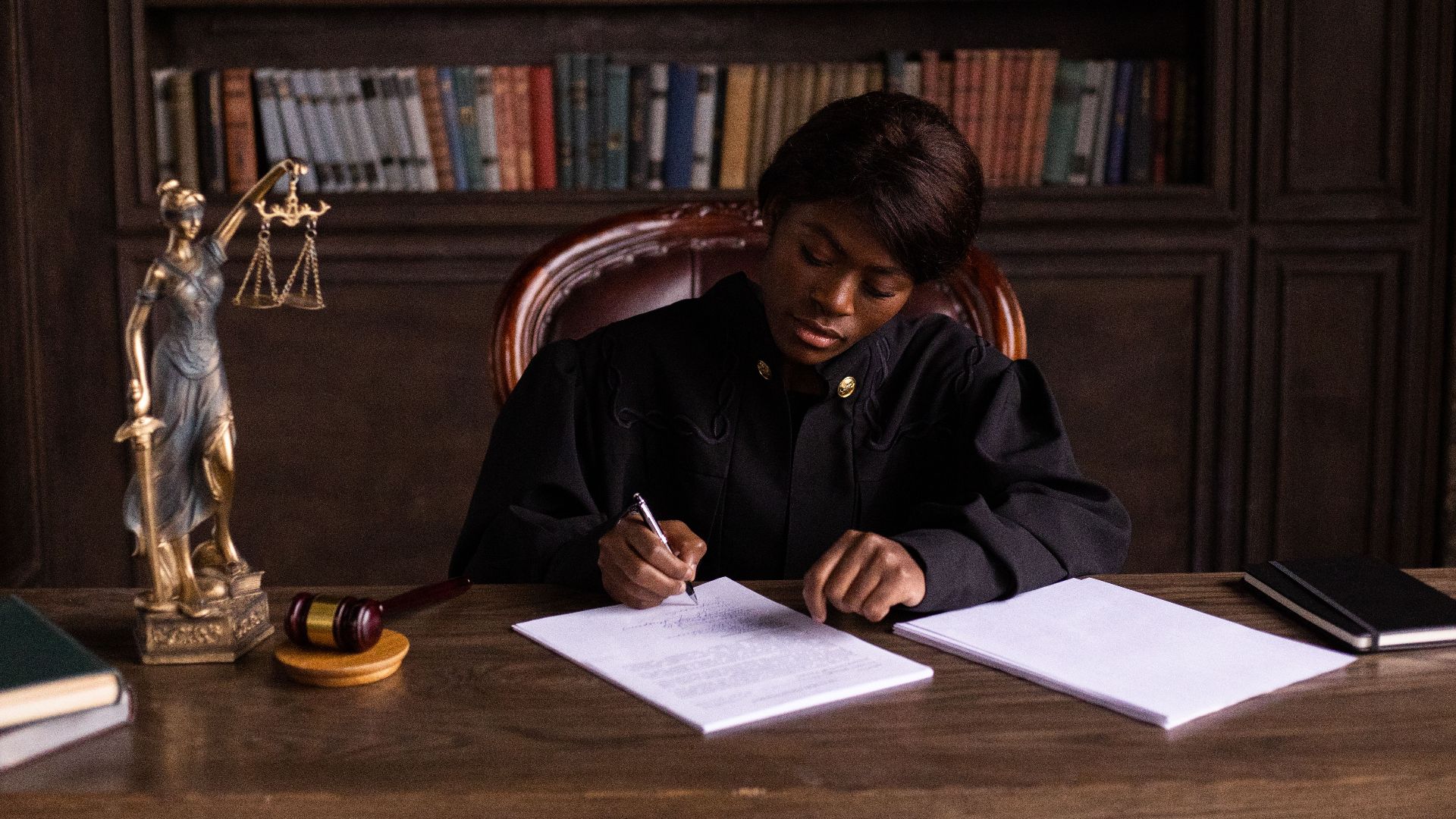 A judge in robes writing on a document at a desk in an office library with law books.
