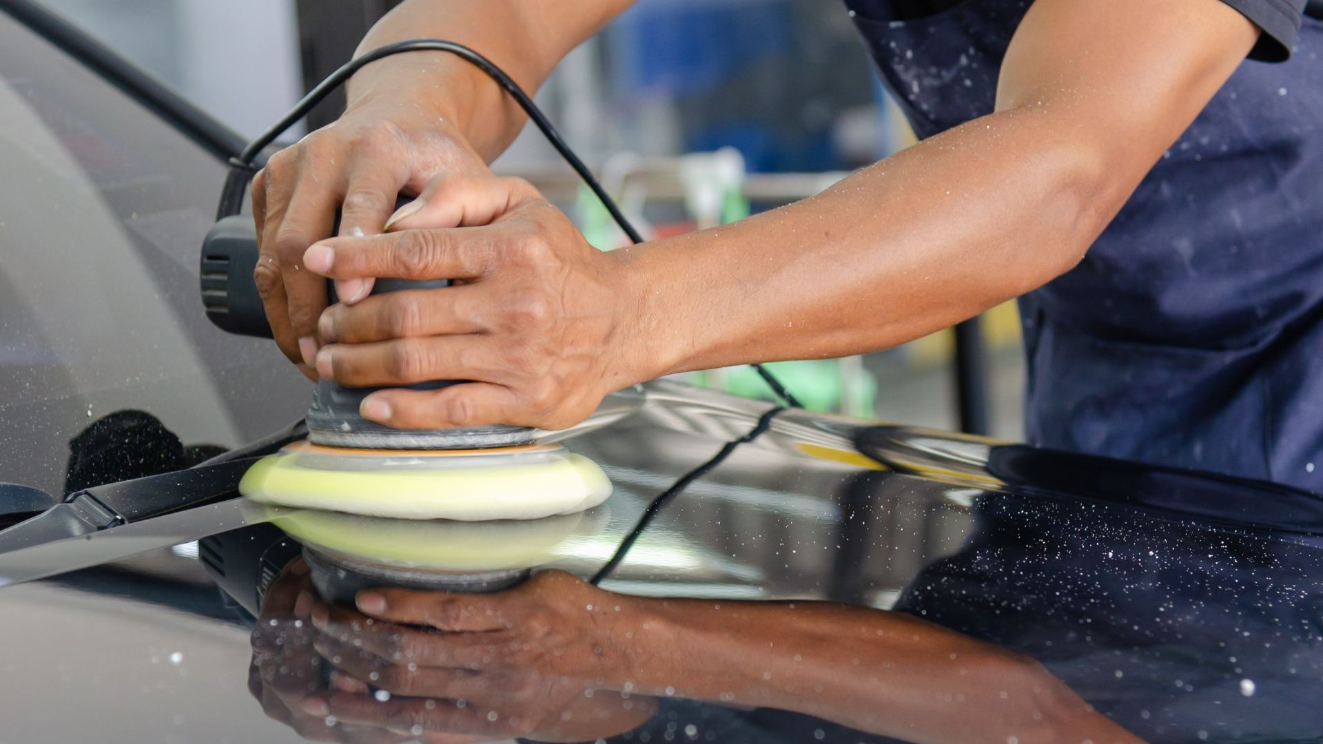 Detailed view of a worker's hands polishing a car's hood using a buffer in an auto workshop.