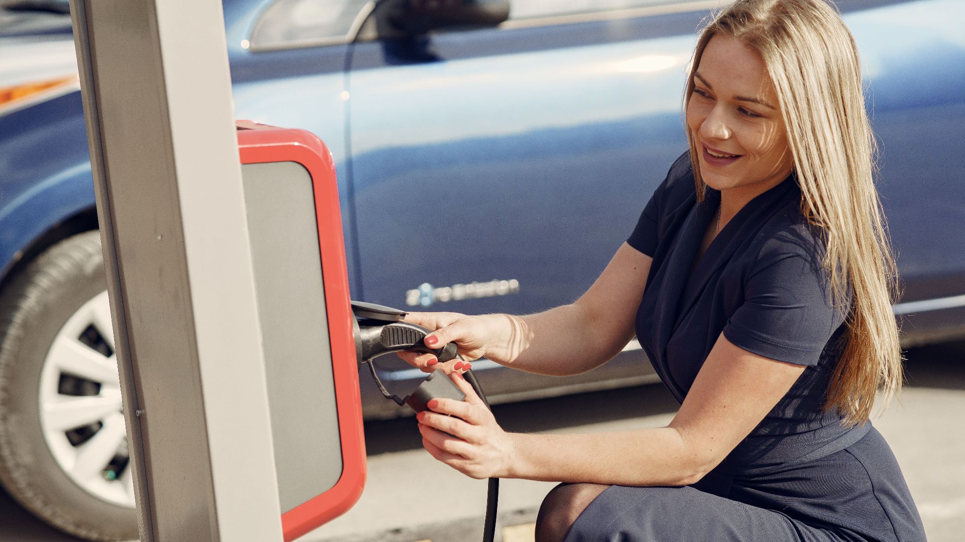 Side view of positive female in casual dress sitting near station on street charging electric vehicle for continuation trip