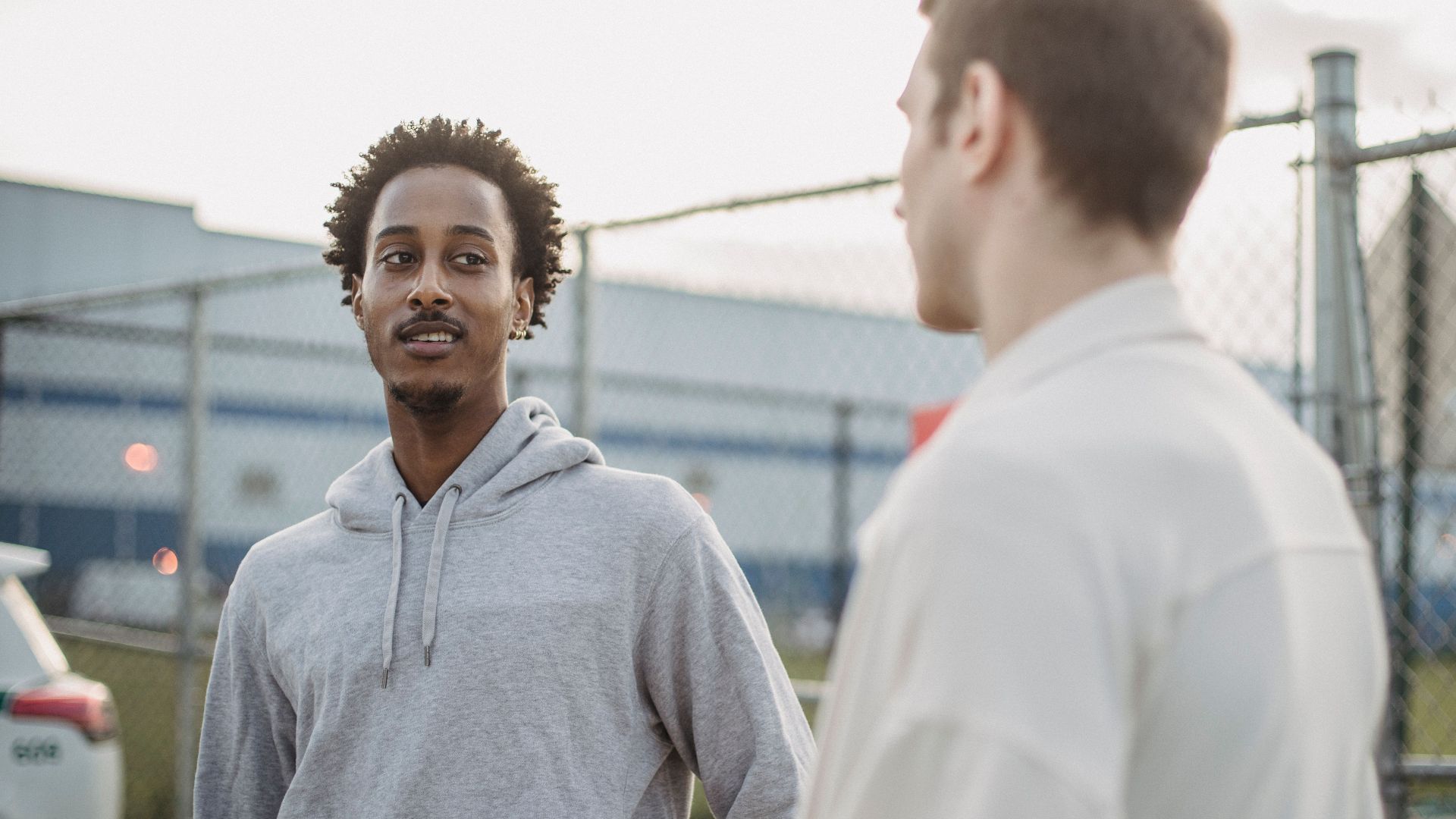 African American male standing with friend against metal grid near car on parking lot