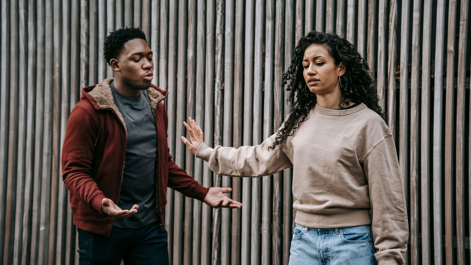Insulted ethnic girlfriend with outstretched arm and irritated African American boyfriend having quarrel near wooden wall on street during breakup