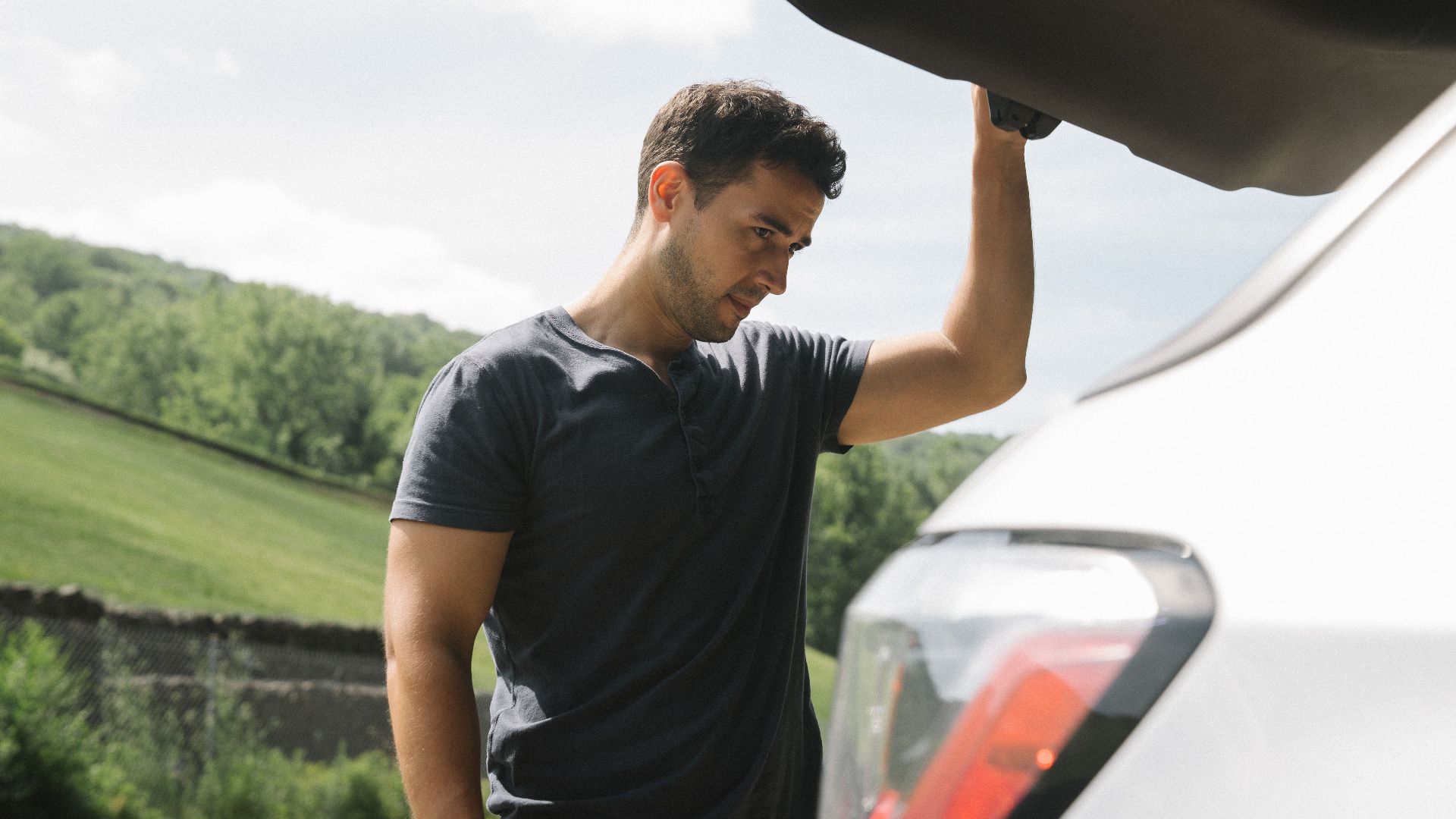 A man examining the open trunk of a car under clear skies with greenery in the background.