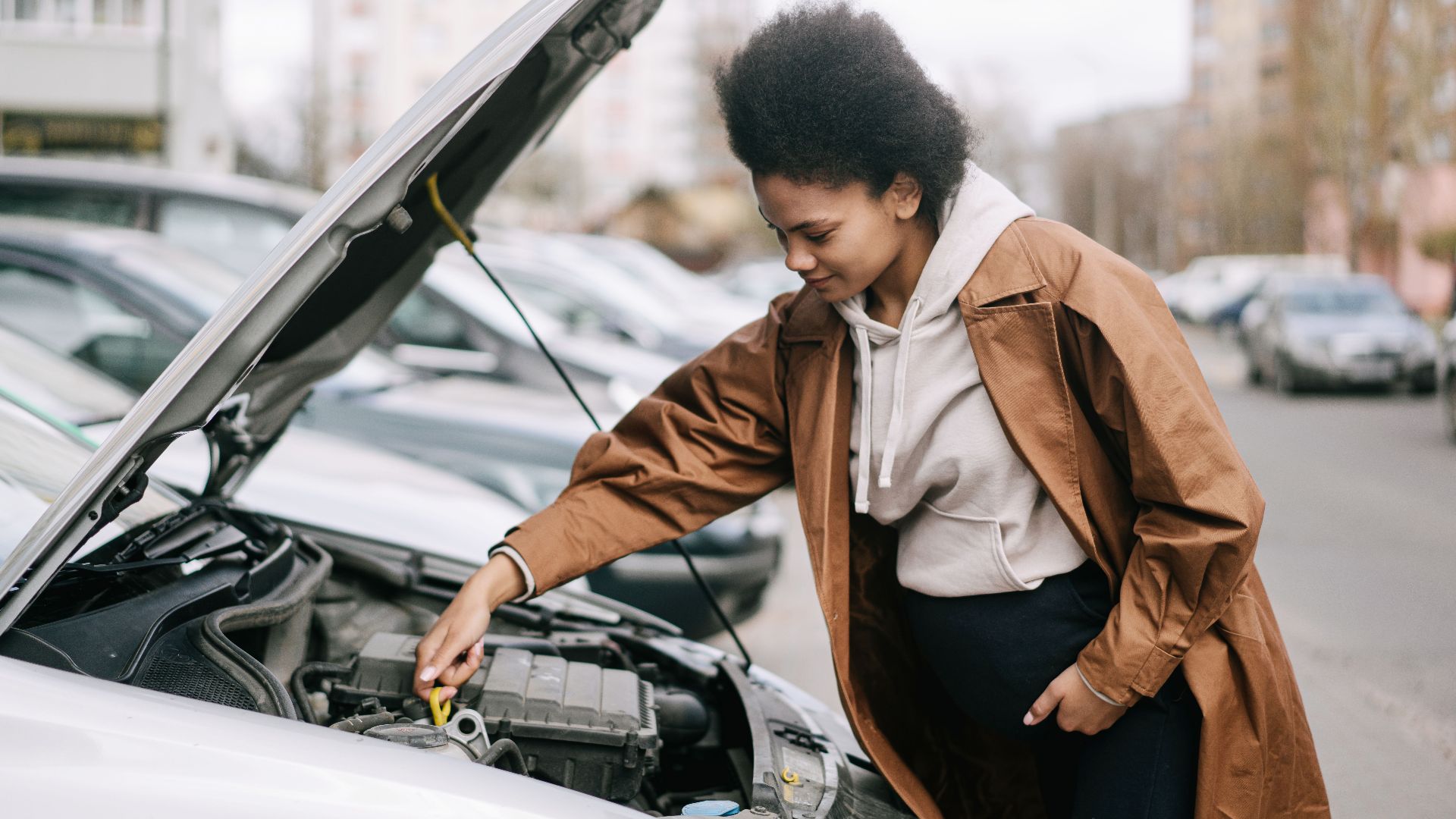 A pregnant woman inspects a car engine outdoors, showcasing independence and car maintenance skills.