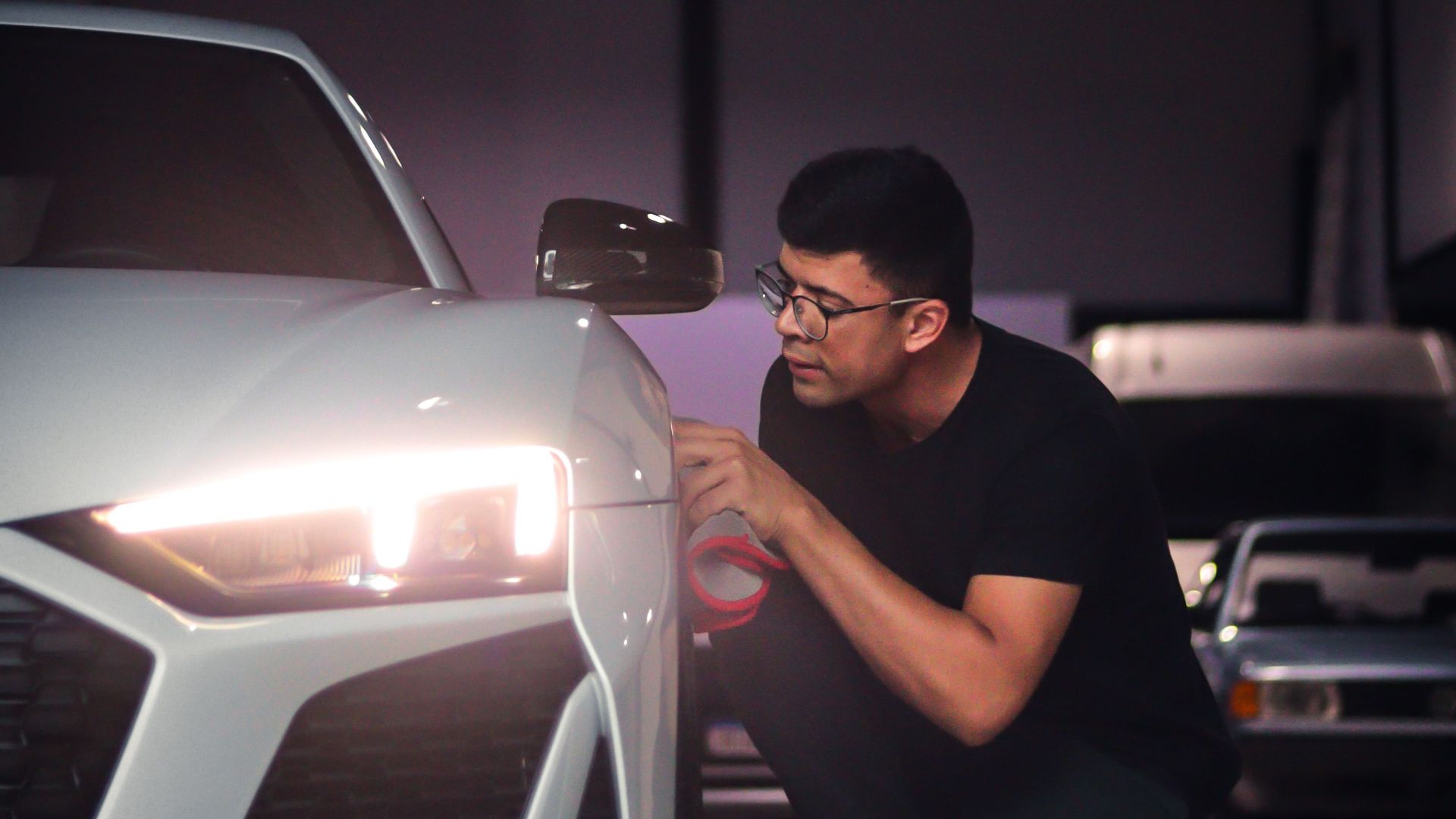 Man kneeling beside a white sports car, inspecting the headlight in a garage.