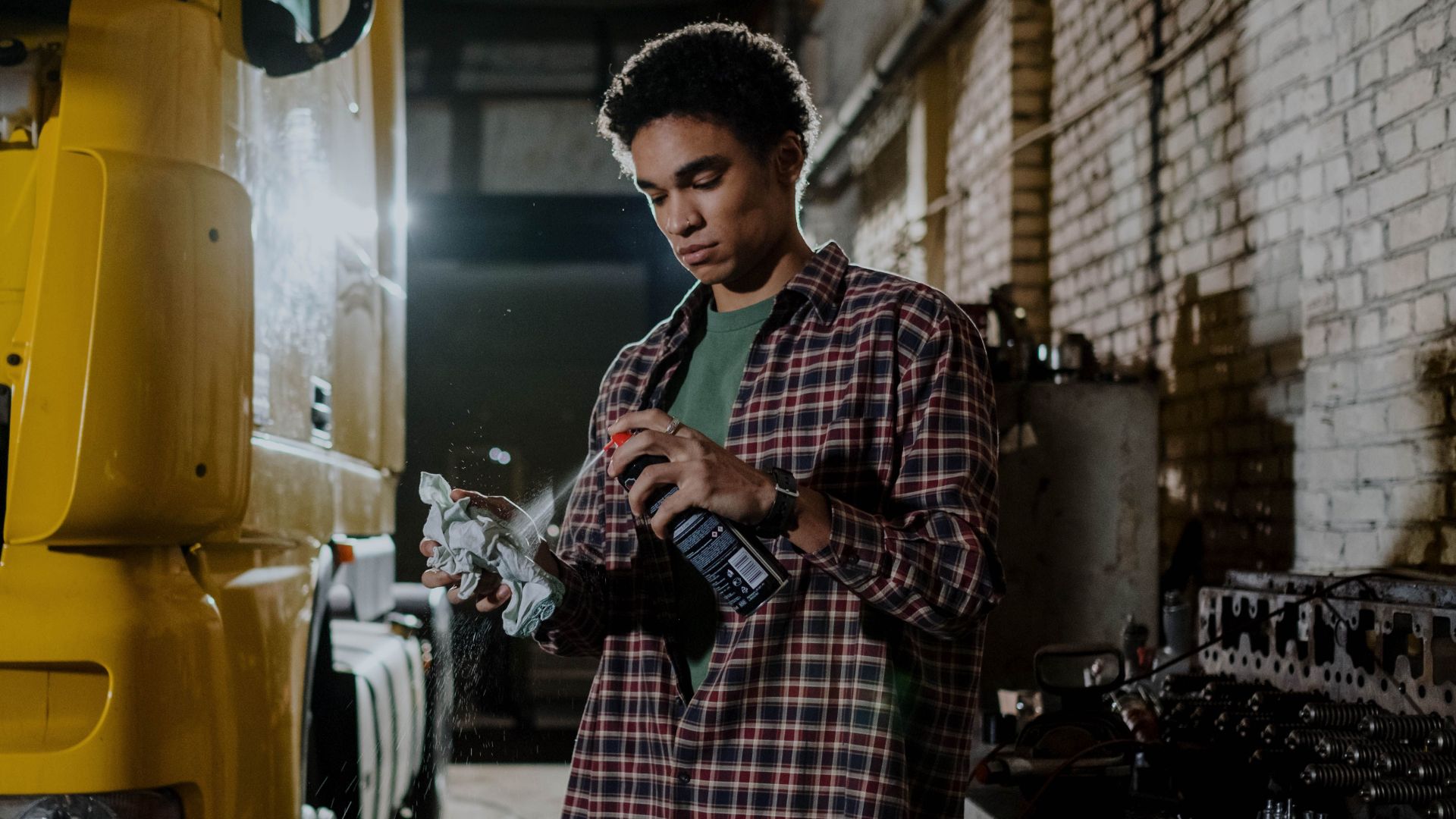 A mechanic in a plaid shirt sprays lubricant while repairing a truck indoors.