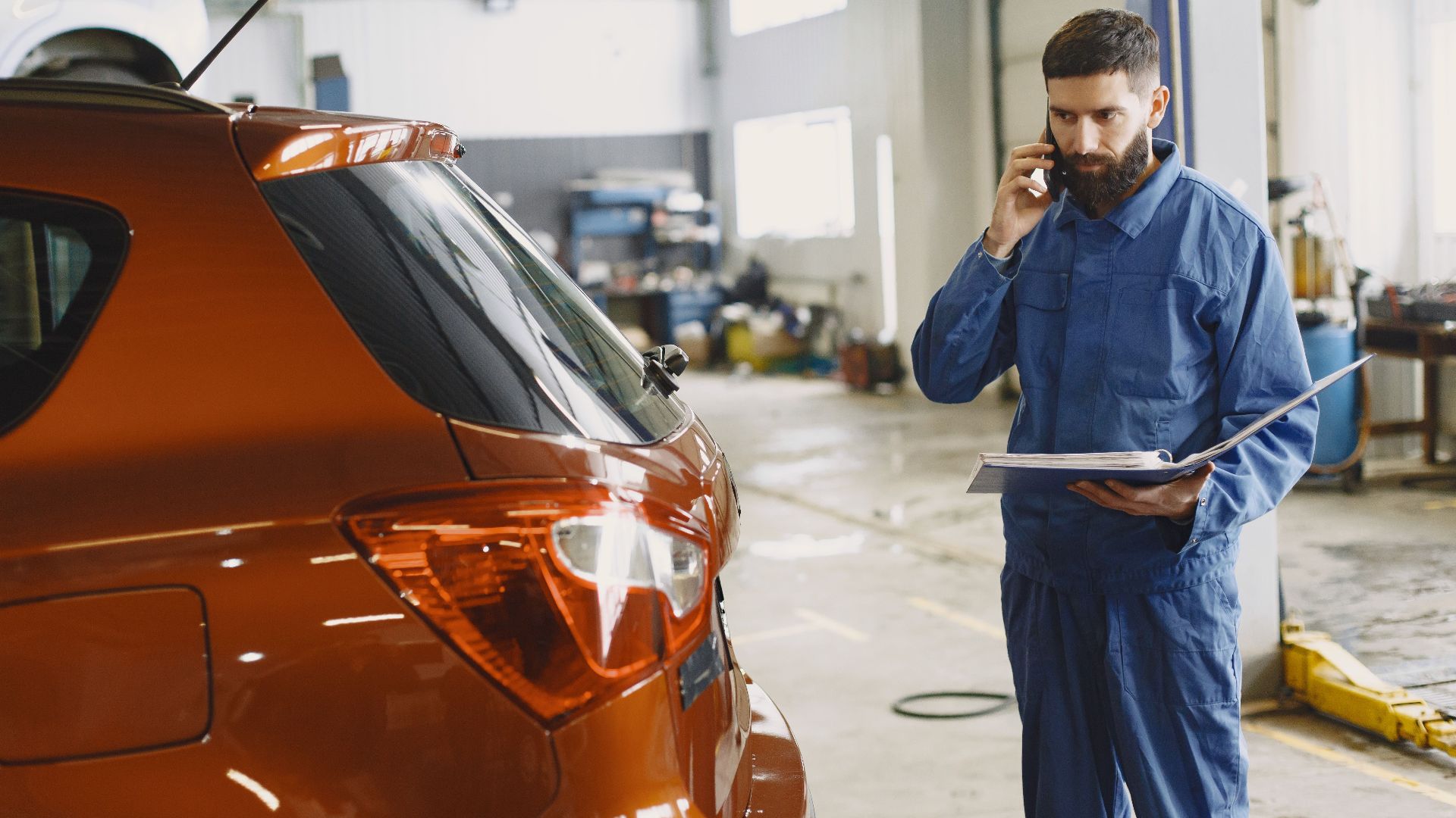 Mechanic in a blue uniform talks on phone beside a red car in an auto repair shop.