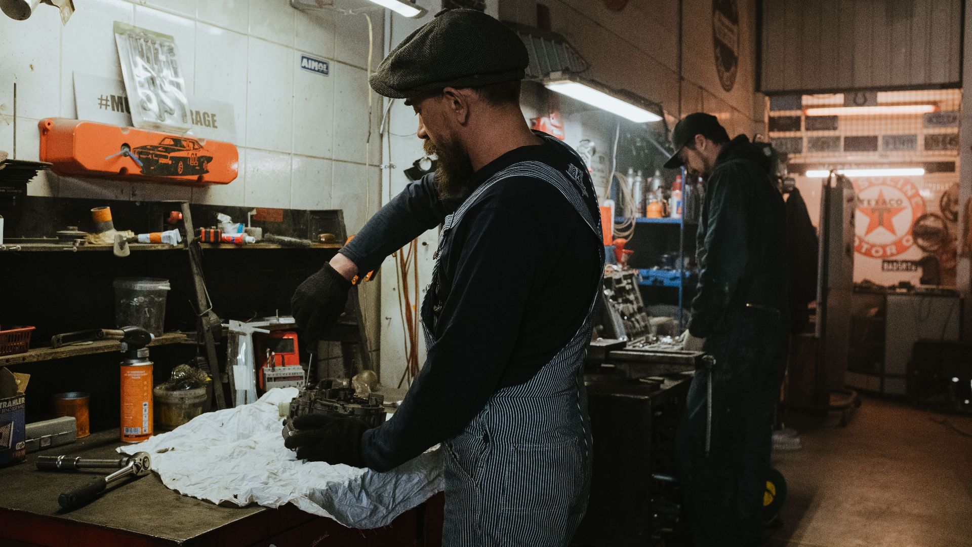 Two auto mechanics engaged in vehicle repair work inside a dimly lit garage.
