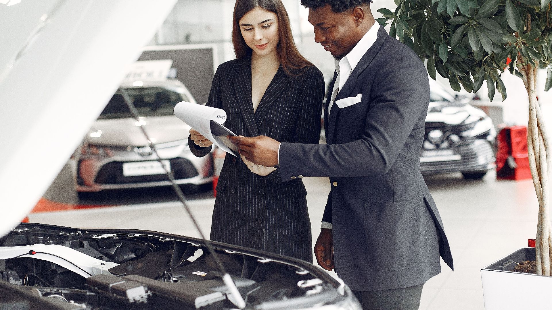 Confident smiling successful black businessman in classy formal suit standing near open motor hood in car showroom and discussing car characteristics with gorgeous female dealer