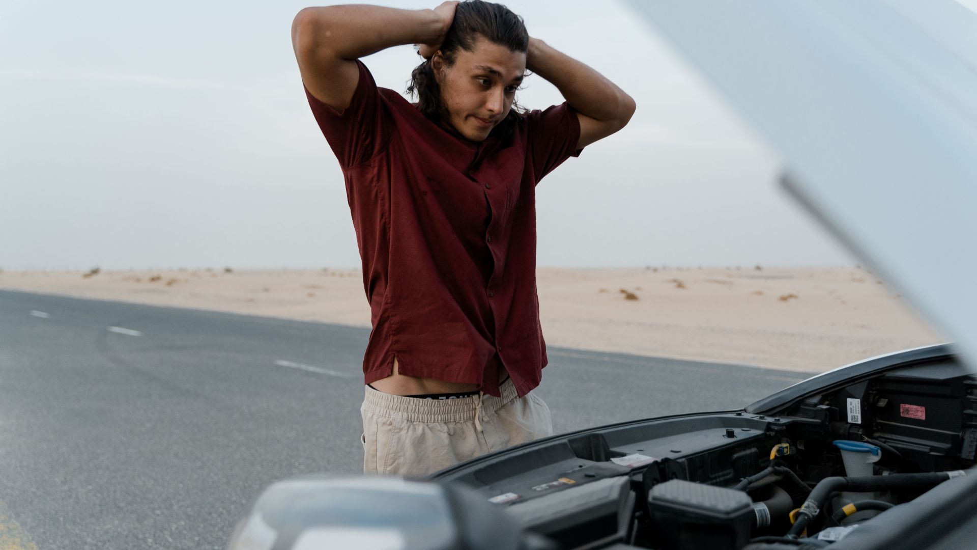 A frustrated man examining a broken car engine on a deserted road, showing stress and concern.