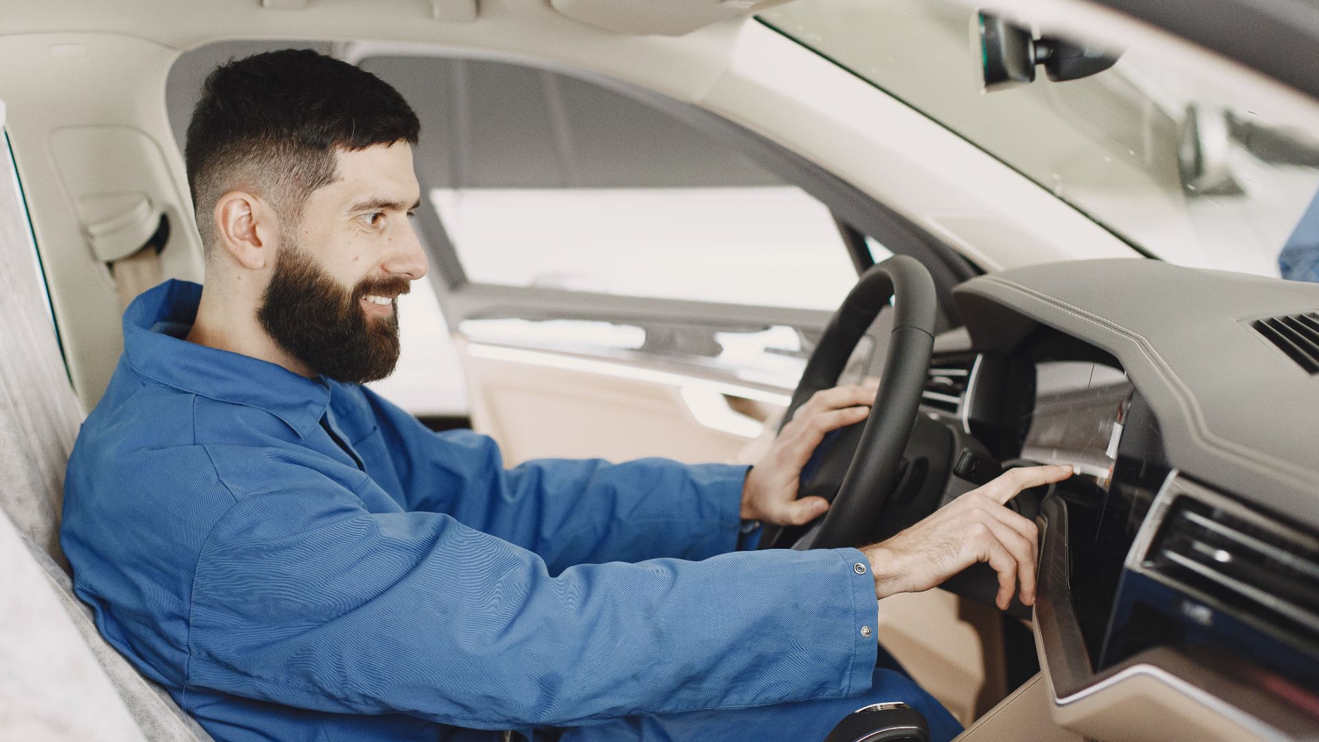 Mechanic in blue coveralls interacts with car dashboard, smiling and focused.