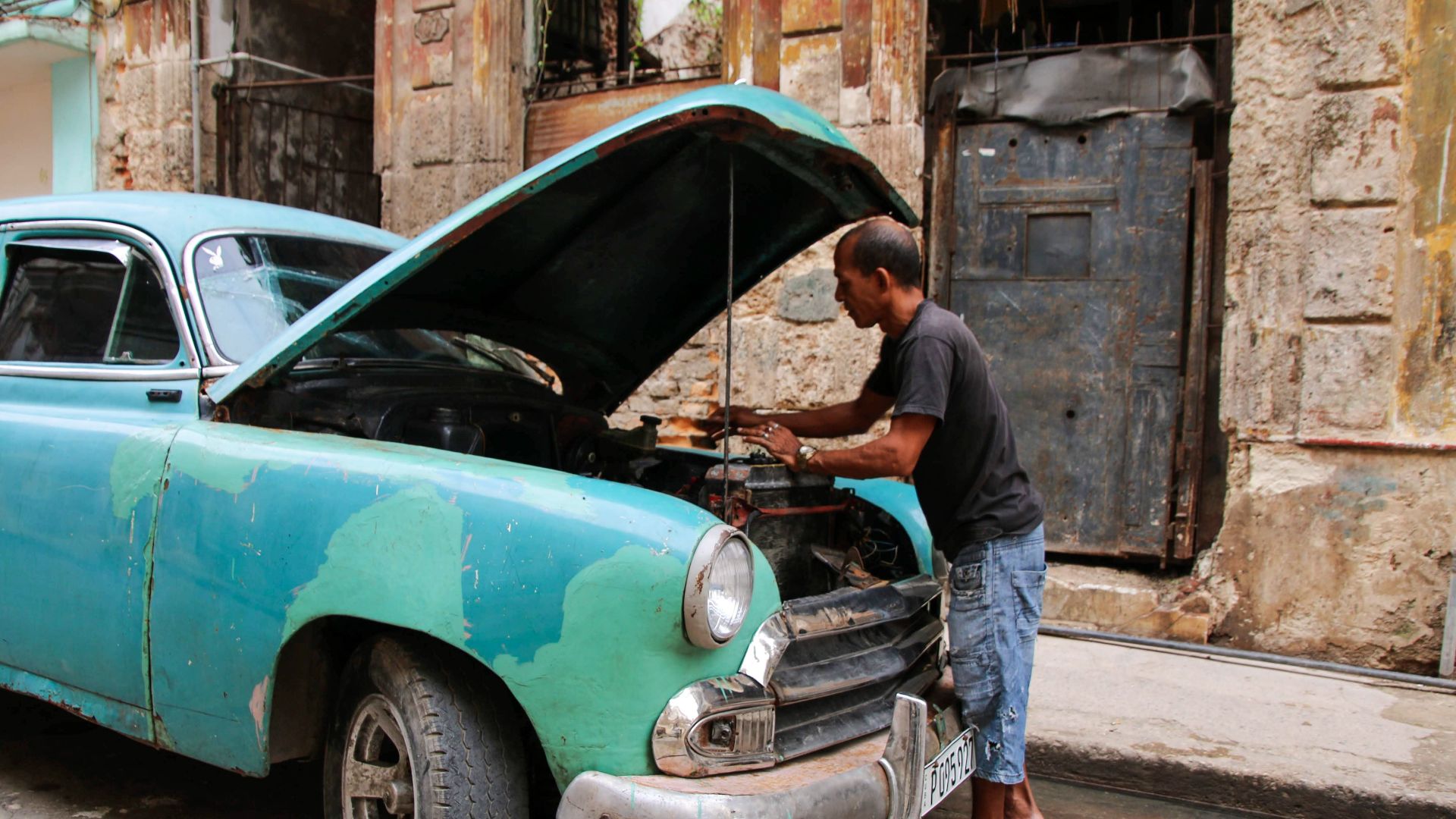 A mechanic repairs an old blue car on a city street, showcasing vintage urban life.