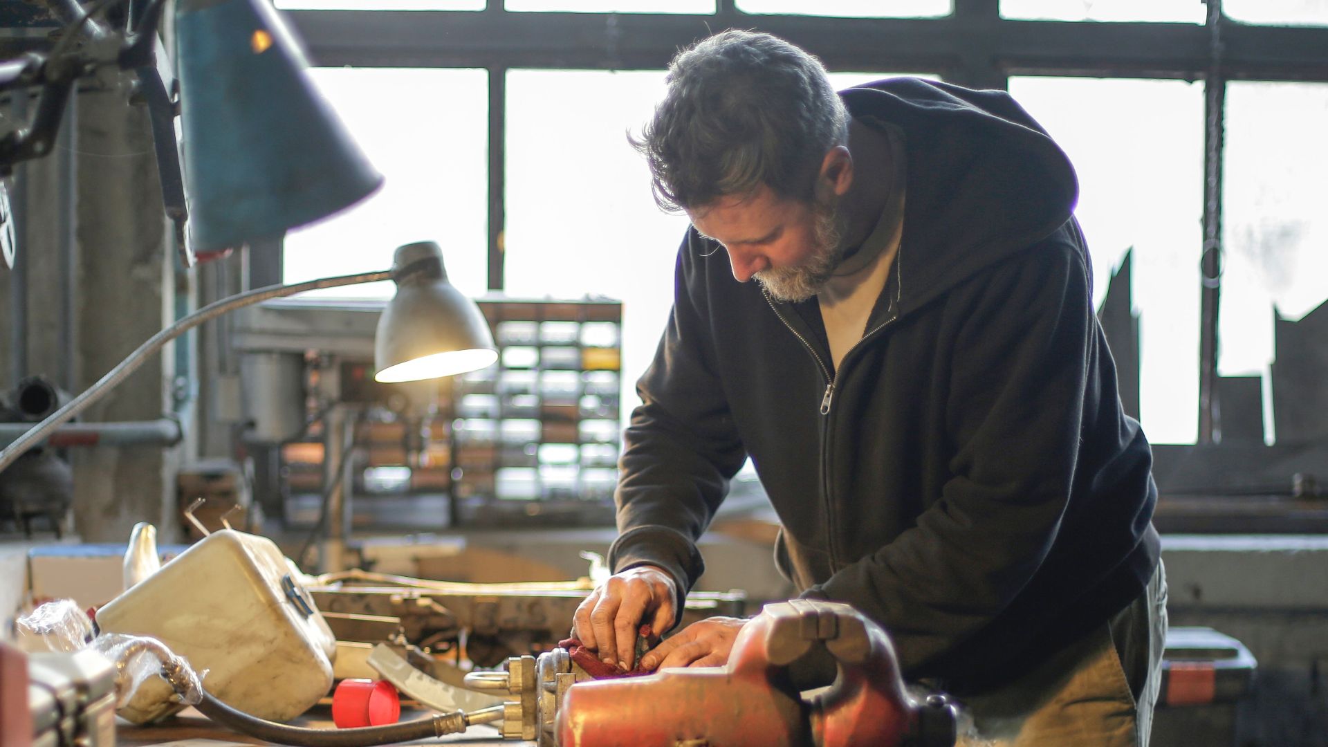 Side view of serious middle aged male in workwear standing near workbench and working with tools and metal details in lamplight in workshop