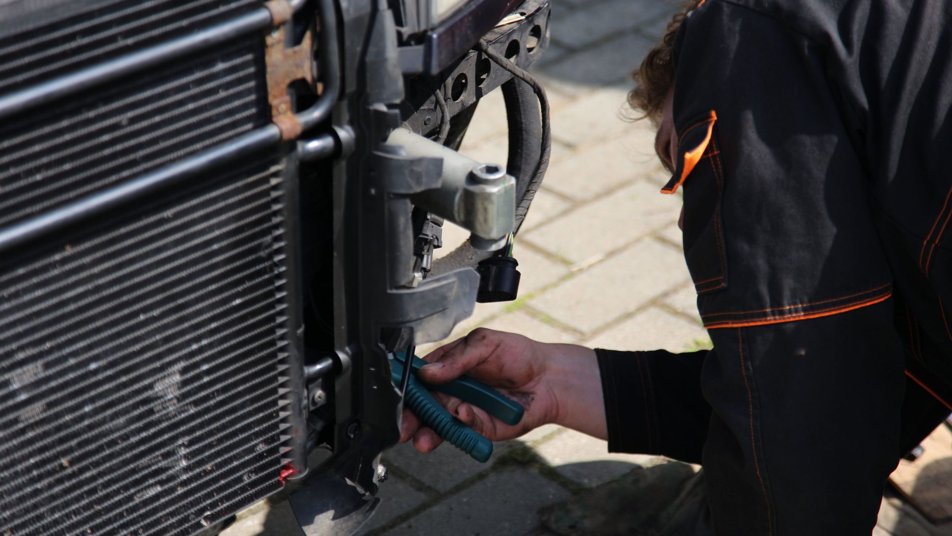 Mechanic working on car repair outdoors using tools, focusing on vehicle radiator area.
