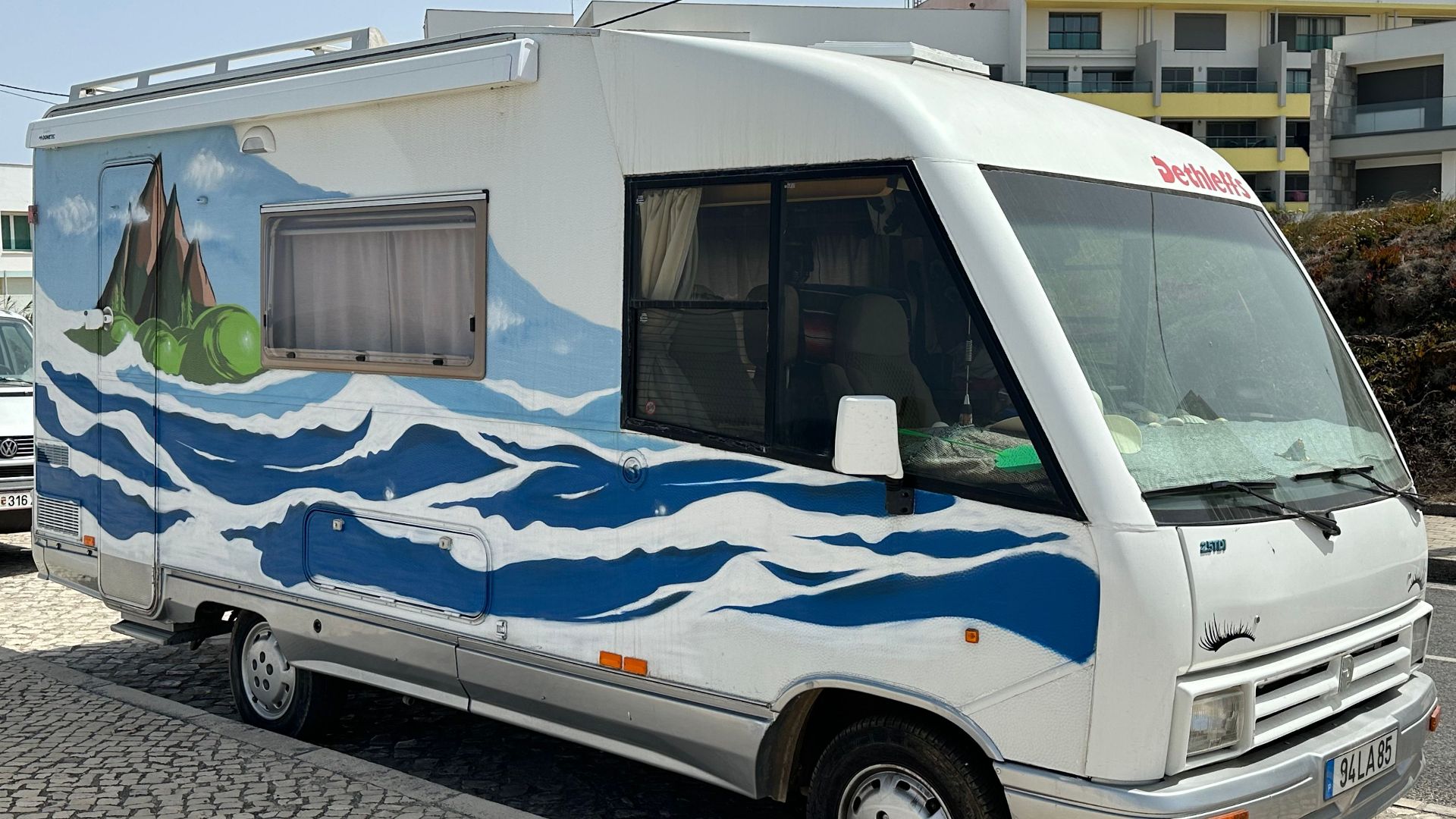 A vibrant camper van with an ocean-inspired mural parked on cobblestone street in sunny outdoors.