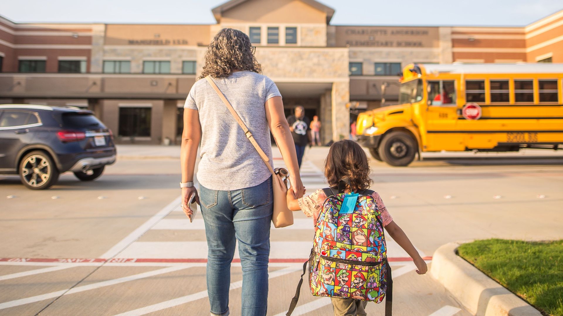 Back view of unrecognizable woman in casual wear holding schoolgirl with backpack by hand while walking on asphalt pavement with marking lines near exterior of building and bus under sky
