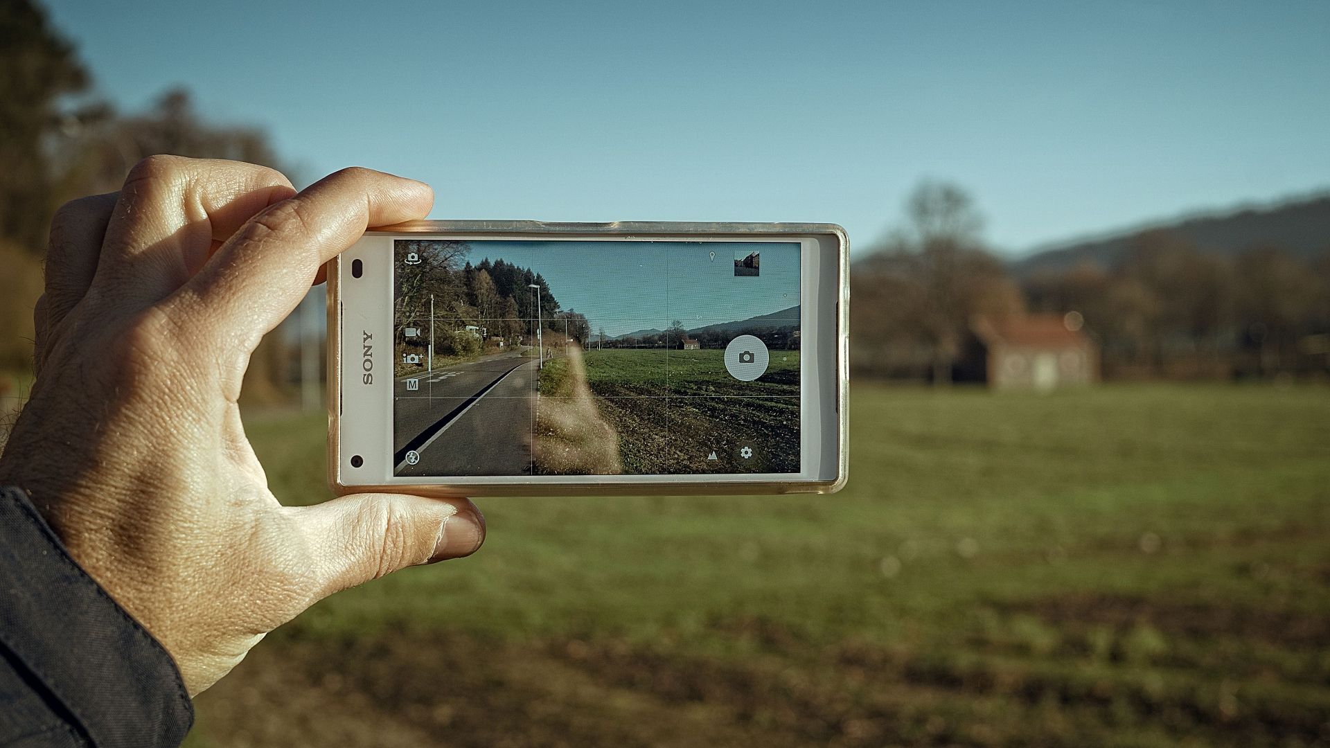 A hand holds a smartphone, capturing a clear image of a scenic countryside road and field.