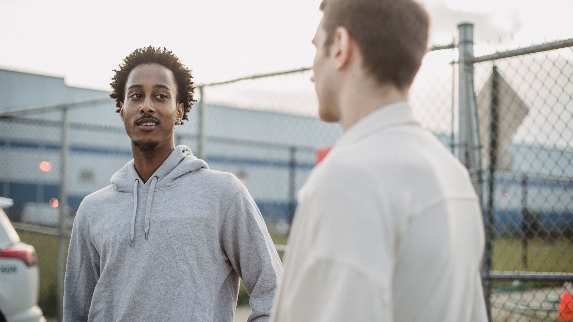 African American male standing with friend against metal grid near car on parking lot