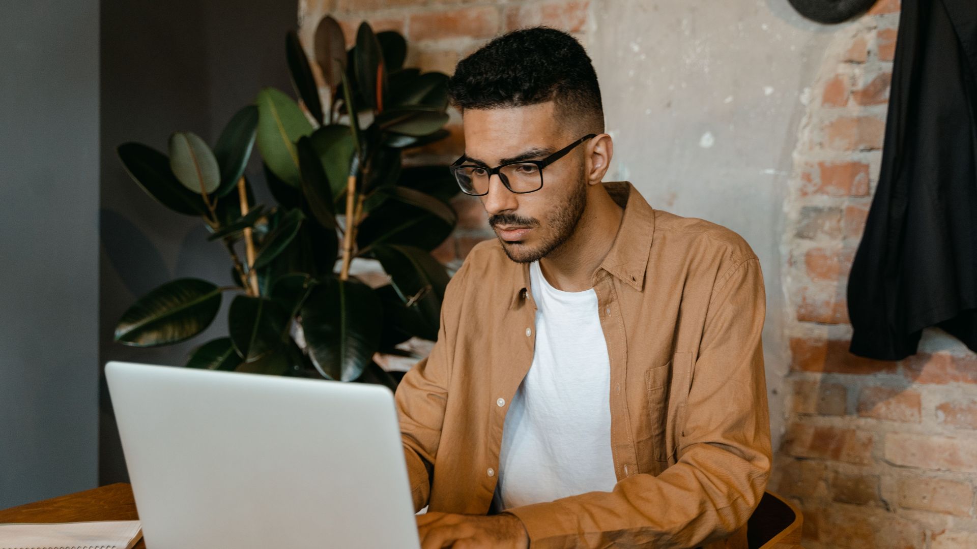 A man in glasses working on a laptop in a cozy cafe with brick walls and plants.