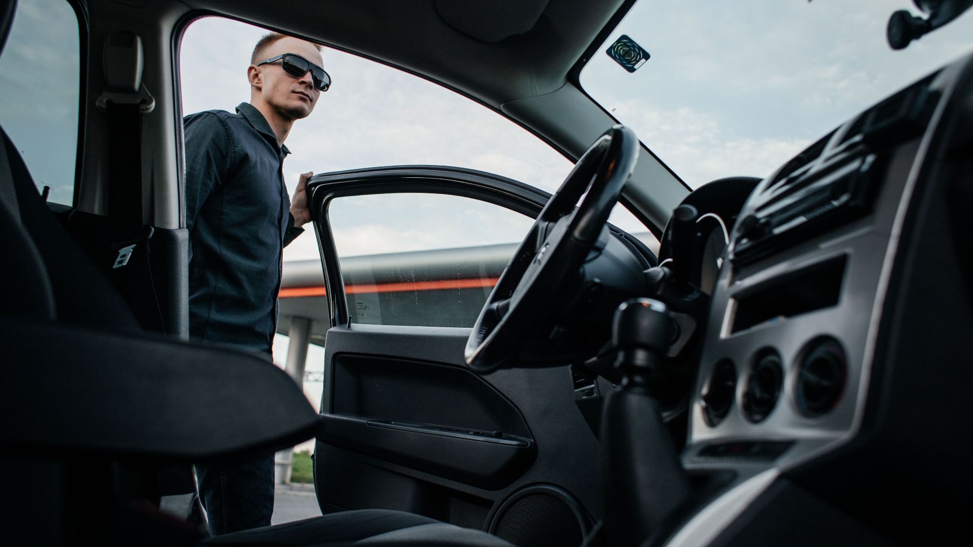 A man in sunglasses stands by an open car door, viewed from the interior.