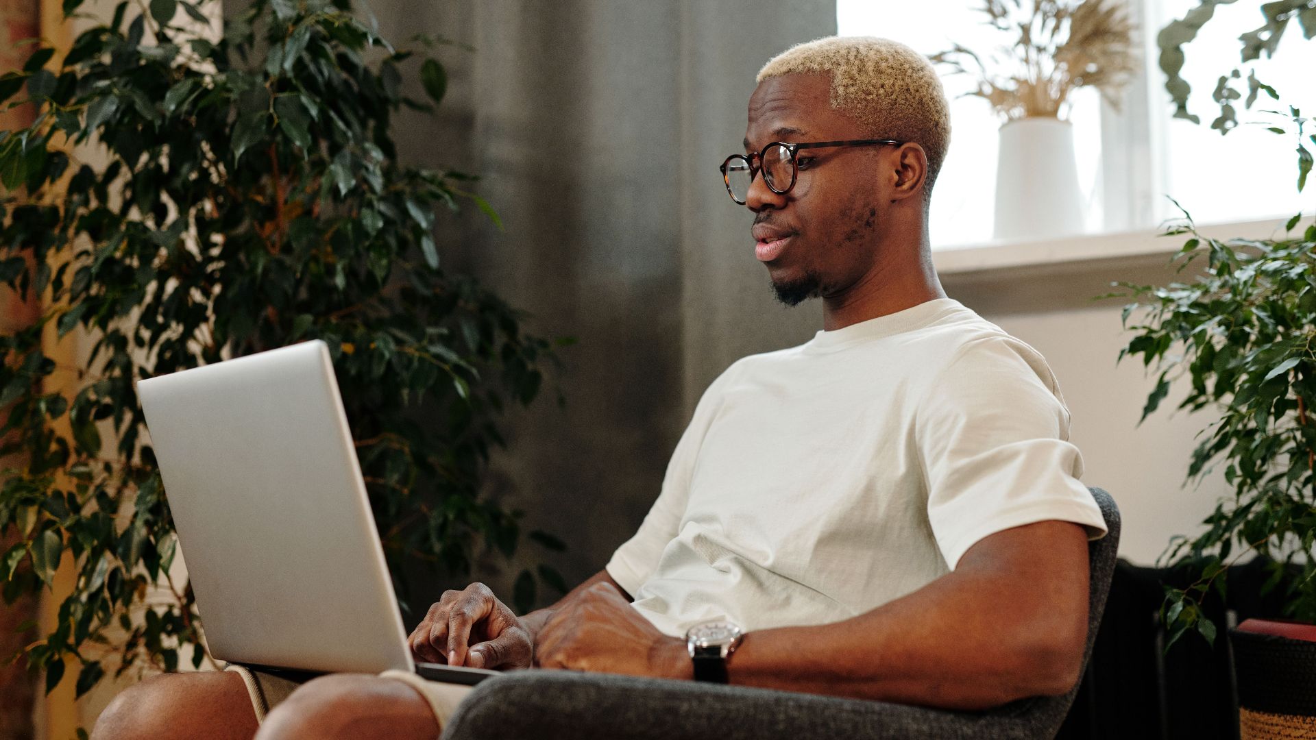 Focused young man with glasses working on a laptop in a cozy indoor space surrounded by plants.