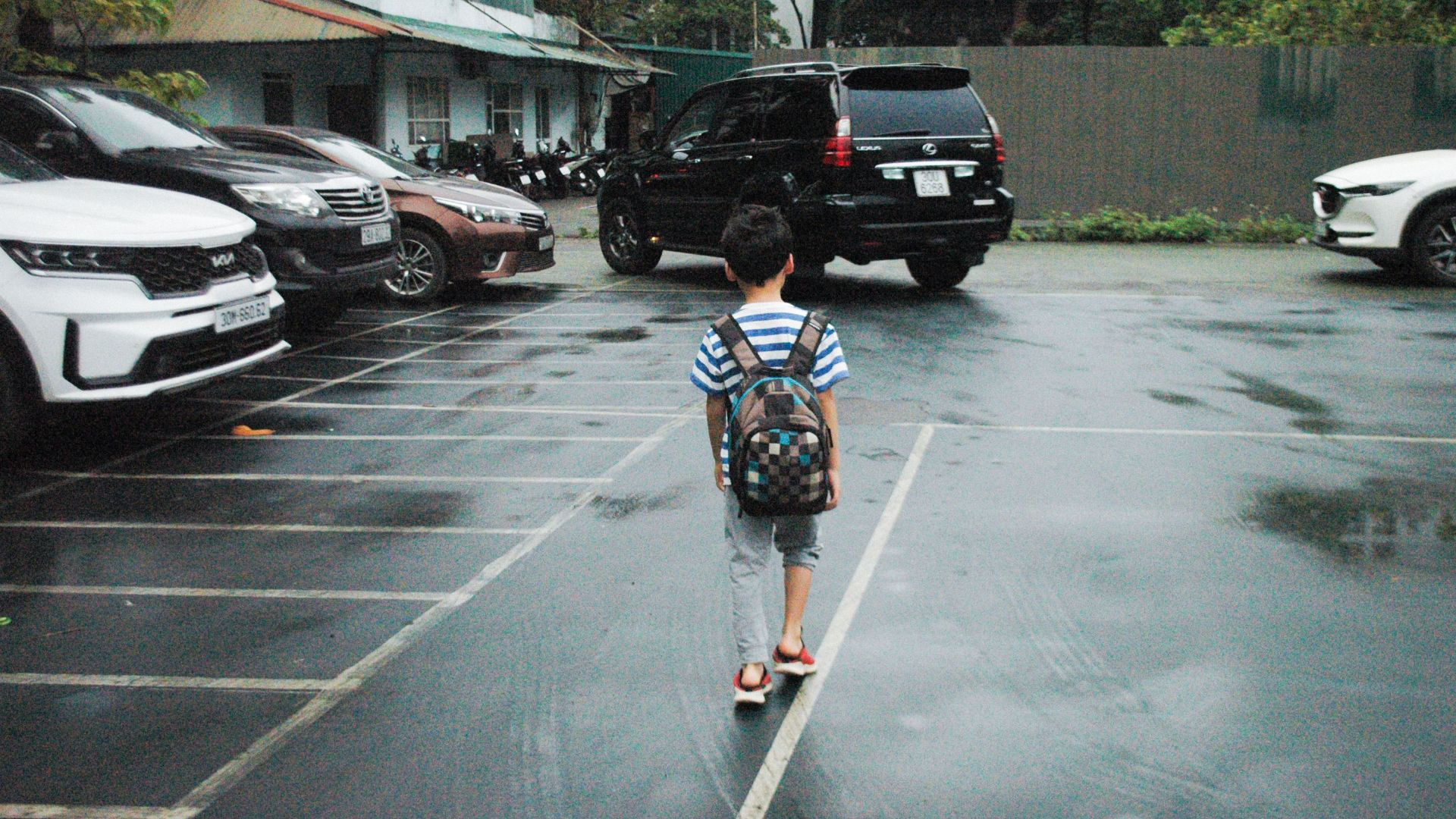 A young boy with a backpack walks through a parking lot, surrounded by cars on a rainy day.