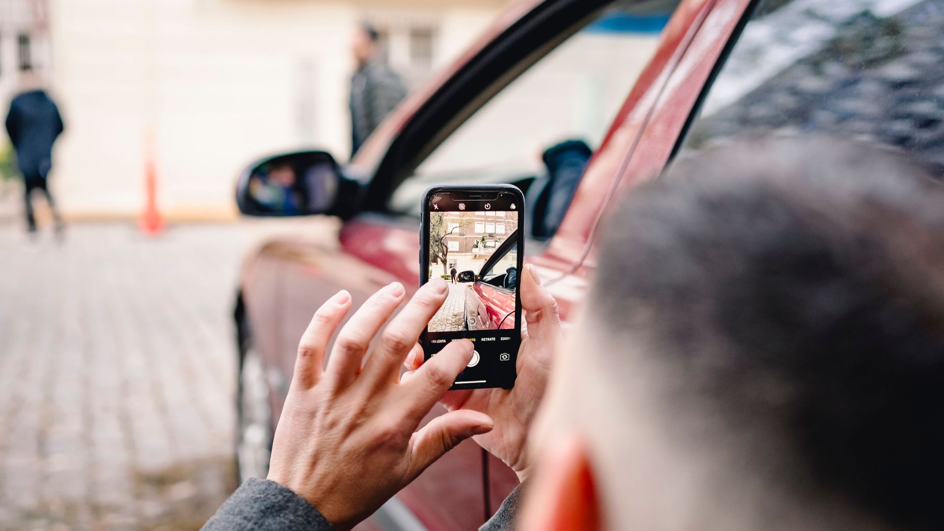 Man using smartphone to take pictures of an urban scene beside a red car.