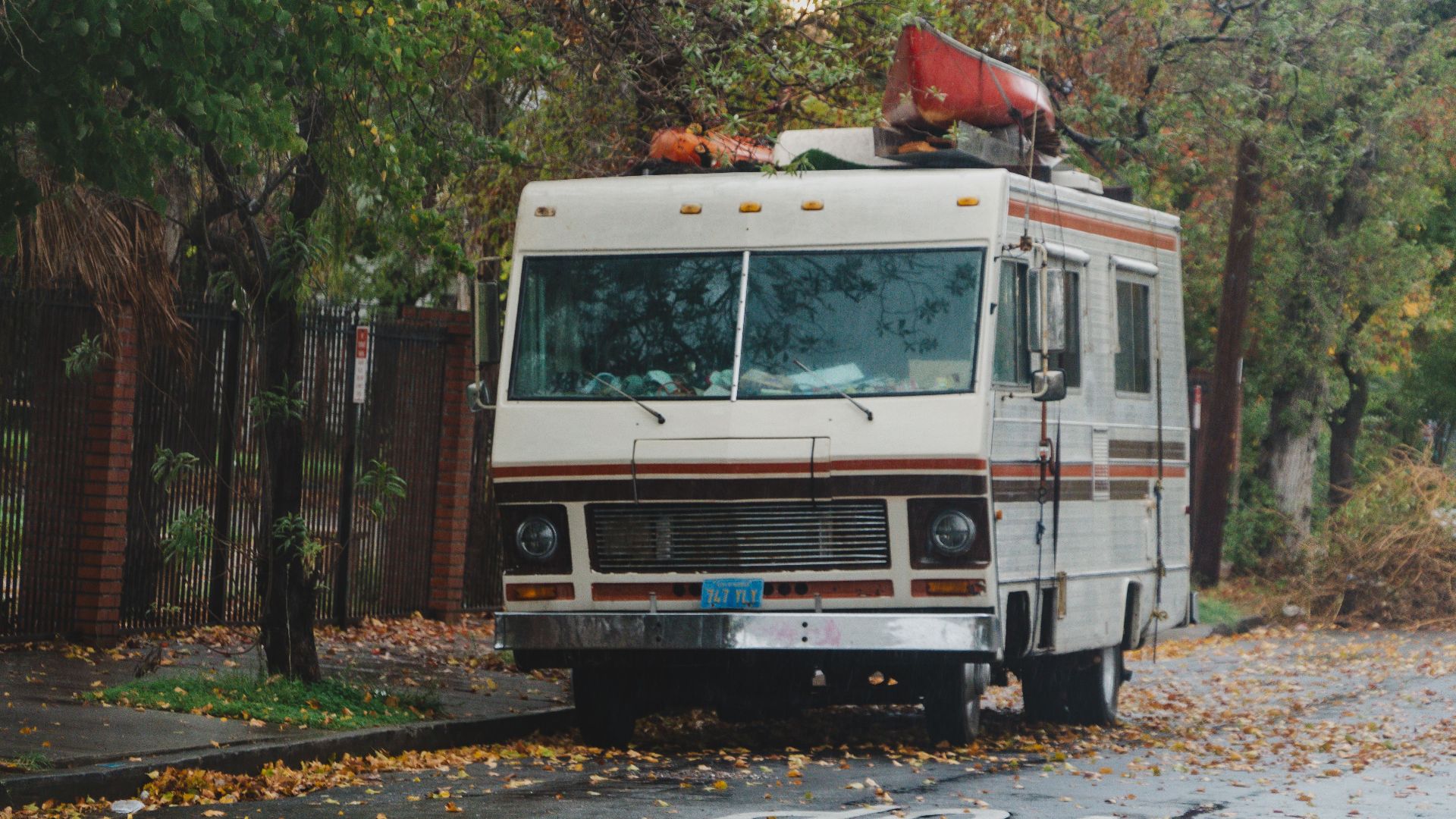 A vintage RV with a red canoe on top parked on a tree-lined street in an autumn setting.