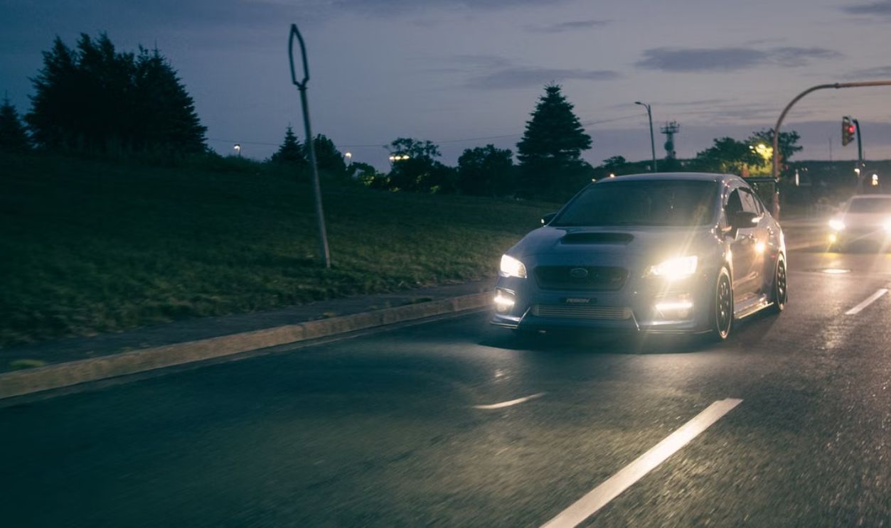 A sleek Subaru WRX drives on a city street at dusk, headlights on, capturing the essence of night travel