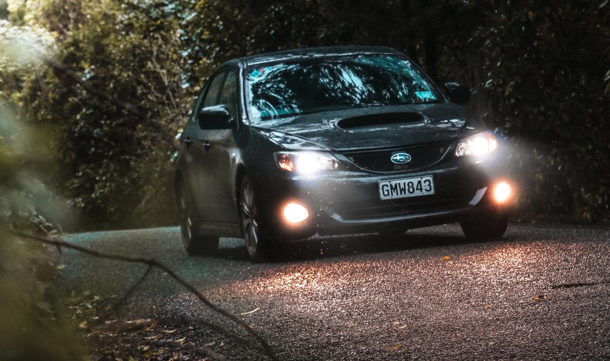 A car with headlights on drives through a winding forest road at dusk, surrounded by trees and foliage