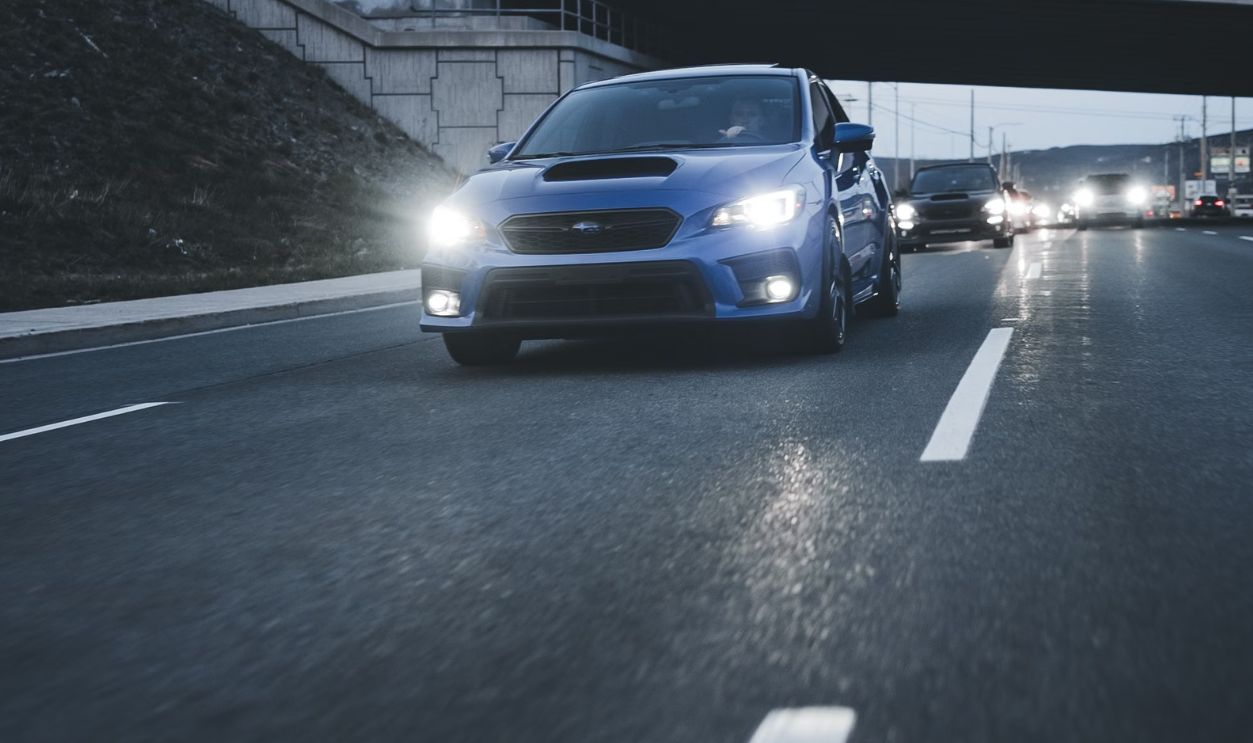 A blue car driving under a bridge on a highway at dusk with headlights illuminating the road ahead