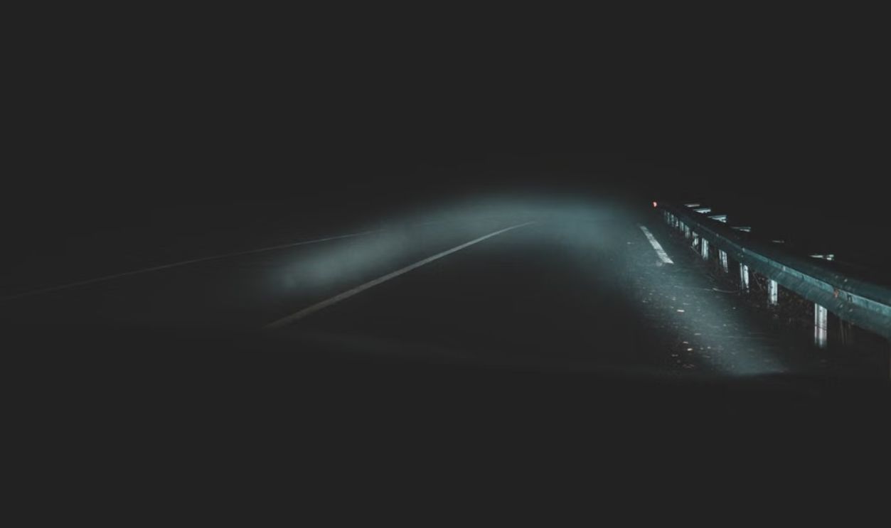 Eerie night road with fog and visible guardrail, illuminated by car headlights, creating a mysterious atmosphere.