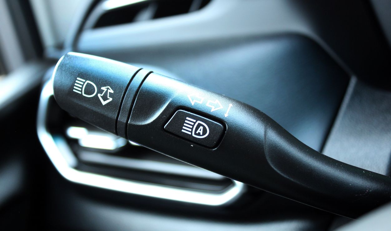 Close-up of the black multifunctional turn signal lever on the steering column. Glossy plastic lever with white icons for headlights, high beams, turn signals, and fog lights, built-in lane change assist button with symbol, matte dashboard trim. Car interior with light switch. Headlight switch; Turn signal lever; Fog light switch.